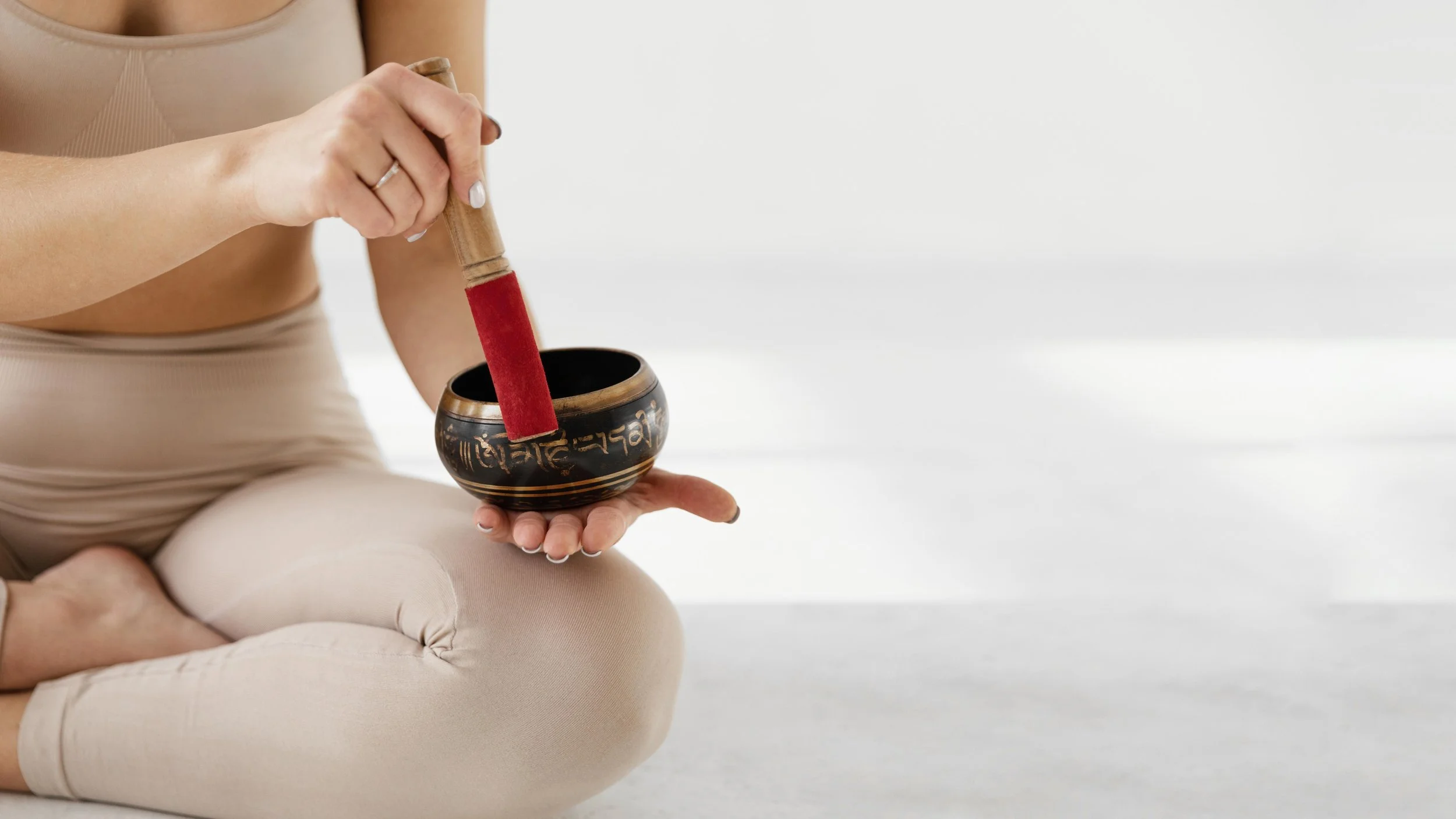 Person sitting cross-legged, holding a Tibetan singing bowl with their left hand and a wooden mallet with red felt on top in their right hand. The person is dressed in beige yoga attire and is seated on a light-colored surface.