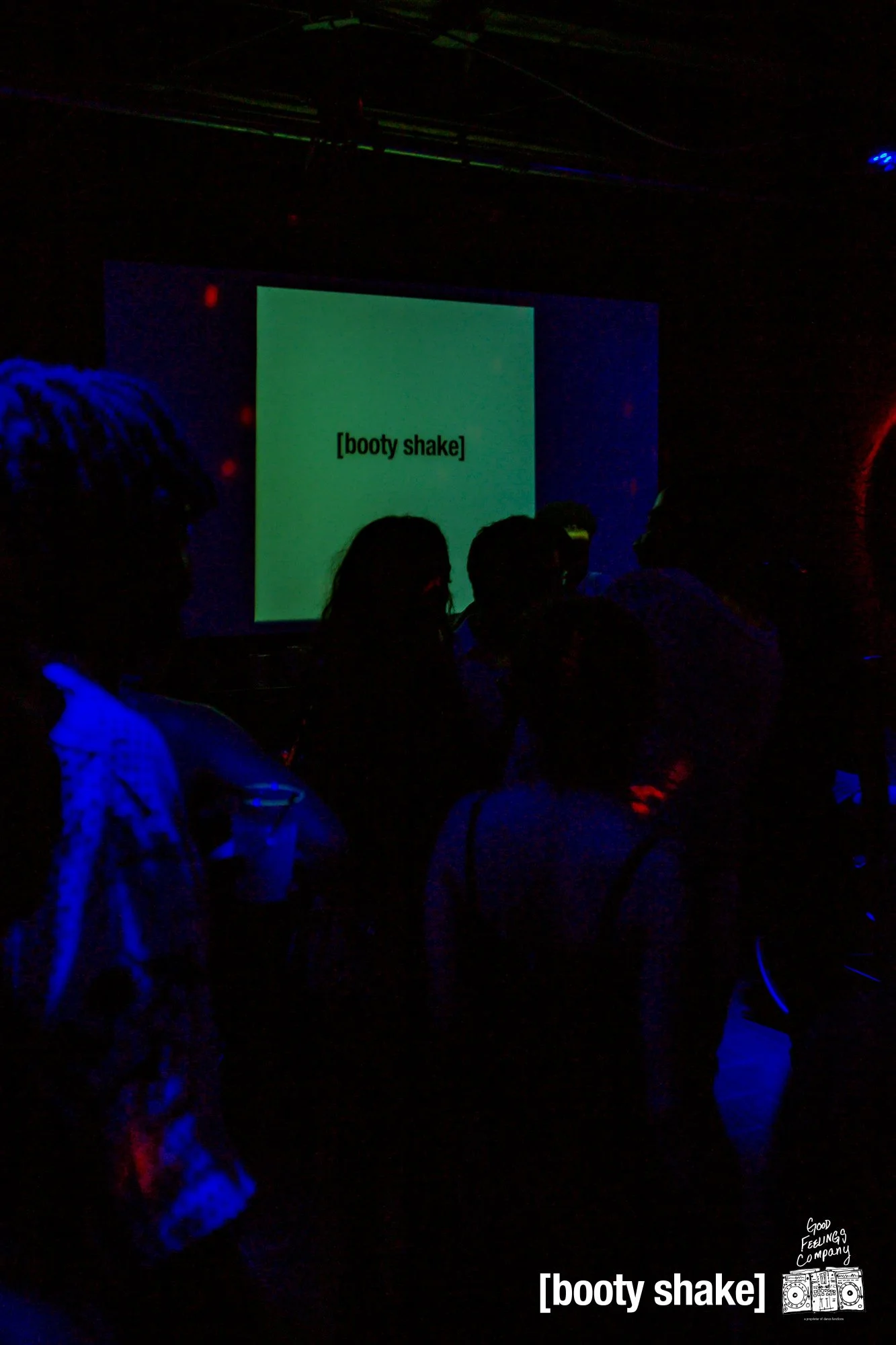 Dark photo of people at a party or club, illuminated by blue and red lights, with a screen displaying the words '[booty shake]' in the background and text '[booty shake]' at the bottom right corner.