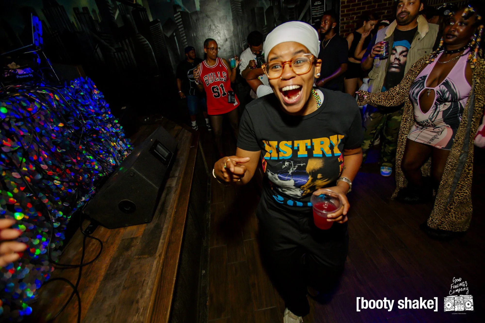 A woman dancing happily at a party or club, holding a red drink, wearing glasses, a headscarf, and a T-shirt with 'HISTORY' written on it, surrounded by other partygoers.