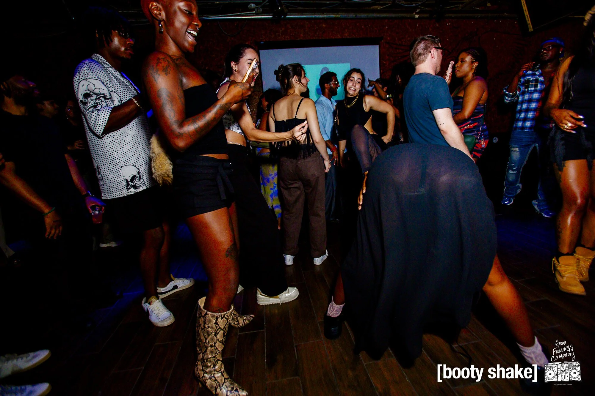 People dancing and socializing at a party with a DJ, some are taking photos or videos, others are dancing closely.