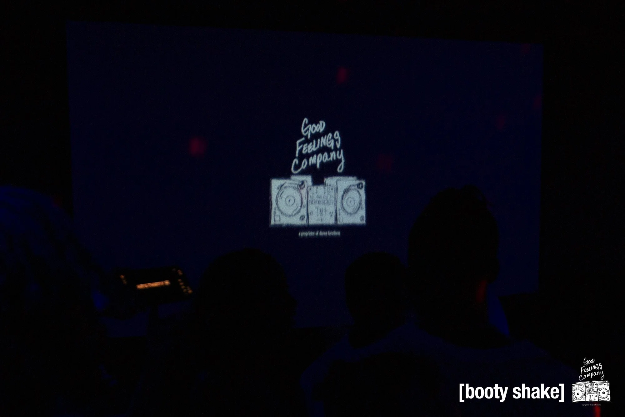 A dark audience area viewing a presentation slide with the text 'Good Feelings Company' and a drawing of a stereo system with the caption 'a portrait of sound sensation'; bottom right corner includes the text '[booty shake]'.