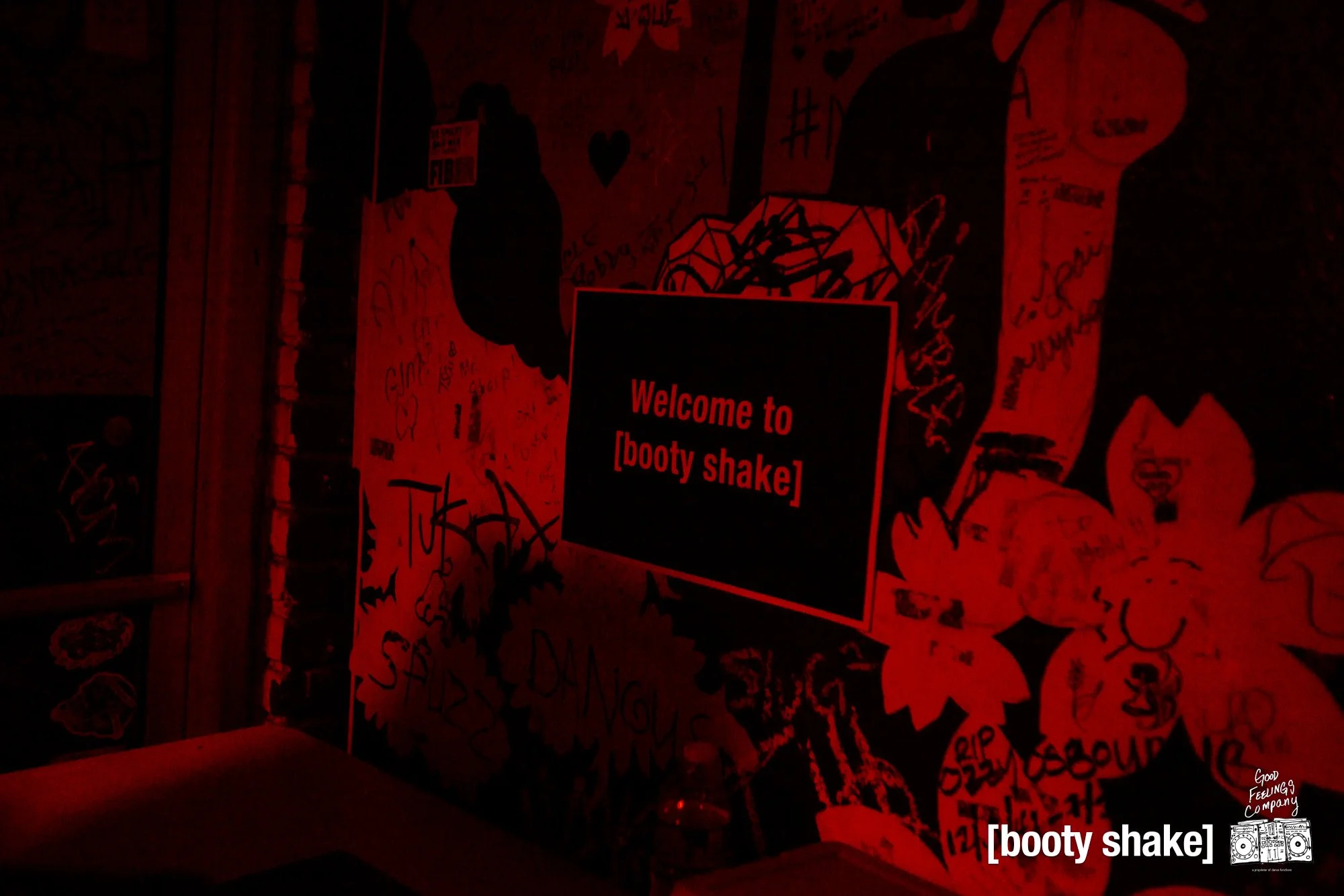 A dimly-lit red and black graffiti-covered wall with a digital sign that reads "Welcome to [booty shake]" and a small logo at the bottom right corner featuring a boombox with the words "Good Feelings Company".