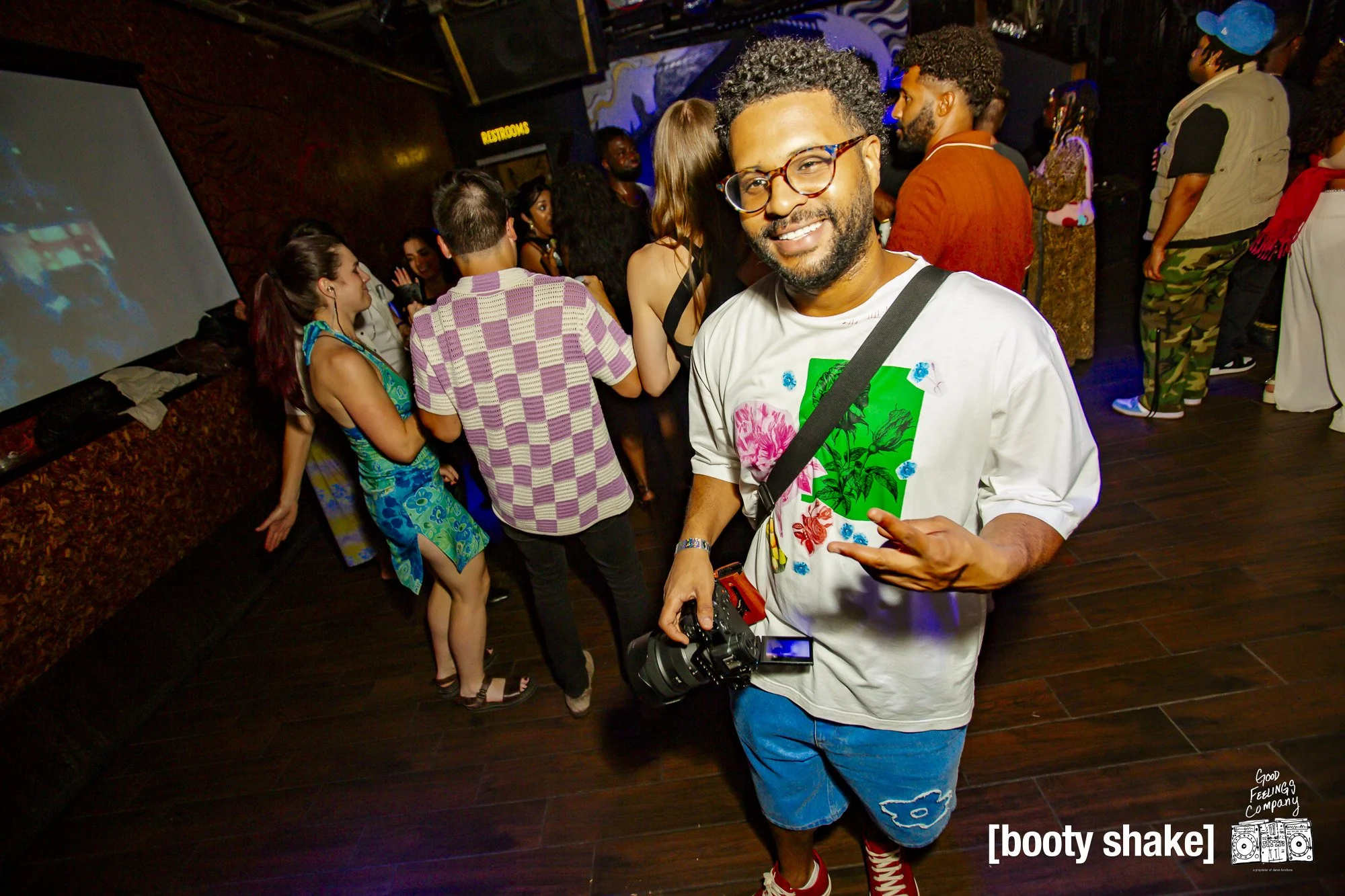 A man smiling and making a gesture with his hand while holding a camera, at a party or nightclub with a diverse group of people dancing and socializing in the background.
