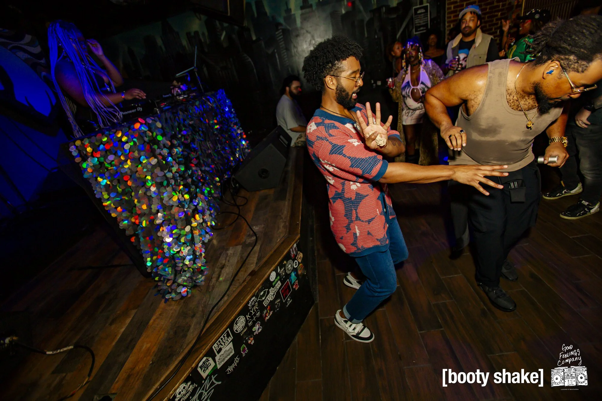 People dancing at a lively party with a DJ booth decorated with colorful lights in the background.
