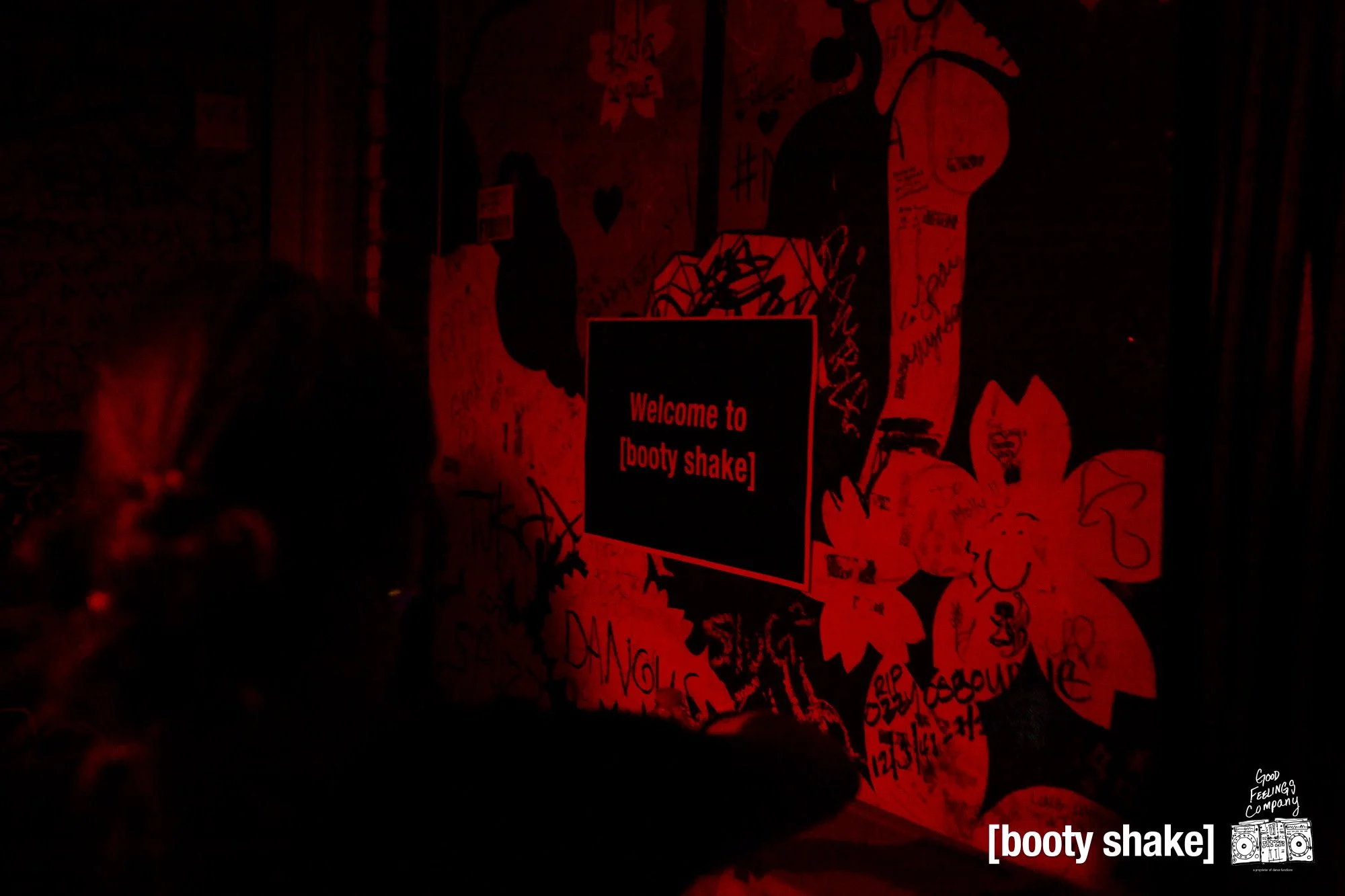 A dimly lit red and black wall with graffiti and a sign that reads 'Welcome to [booty shake]'. There is a person in the foreground and a small logo of a boombox with the text 'Good FEELINGS company' in the bottom right corner.