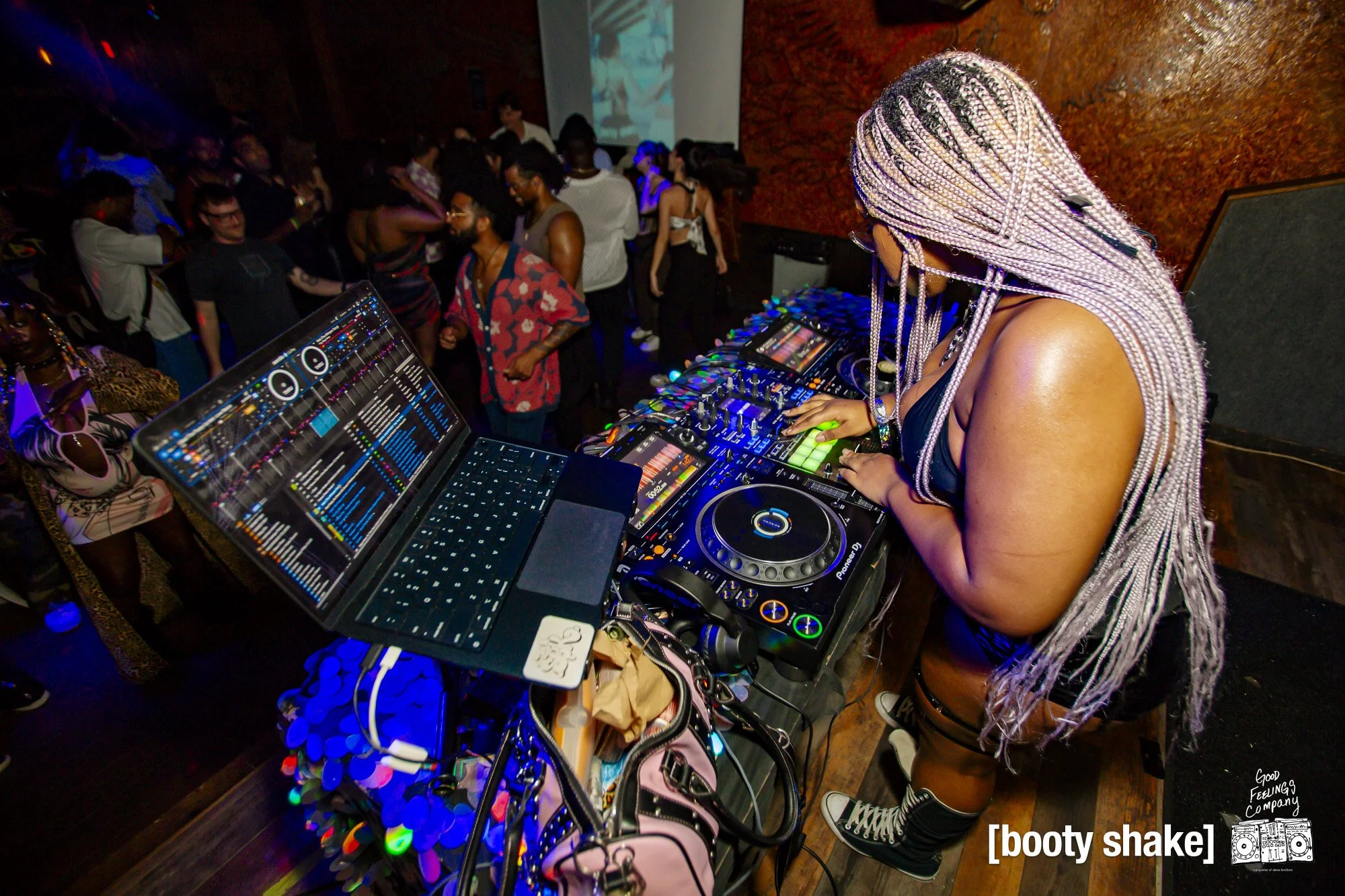 A female DJ with braided hair, wearing a black outfit and sneakers, performs on a DJ console at a nightclub with a crowd dancing in the background.