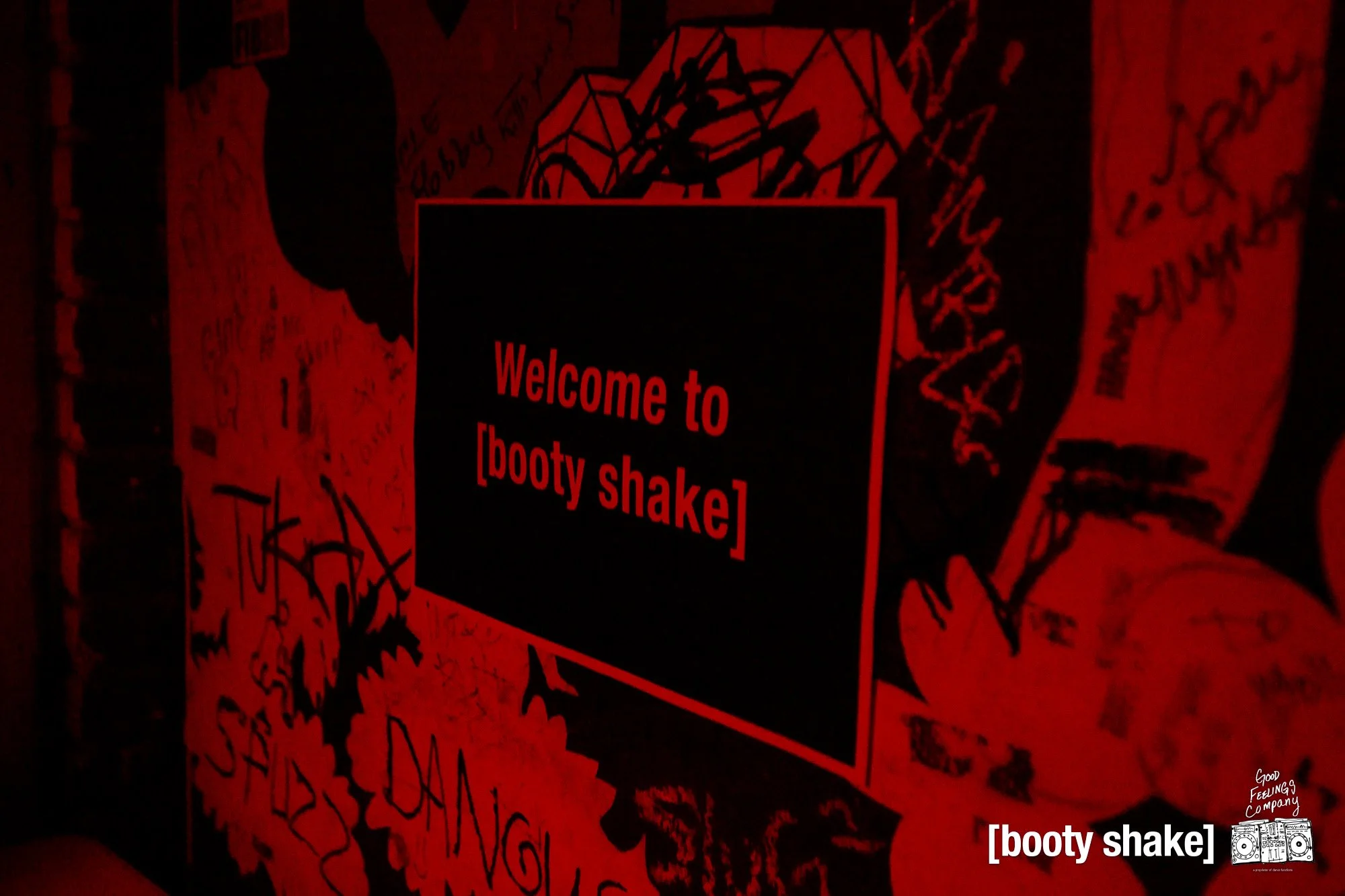 Dark red wall with scribbled writings and drawings, illuminated by red and black lighting, featuring a black square with red text that reads "Welcome to [booty shake]" and white text in the bottom right corner that says "[booty shake]" and a small lo