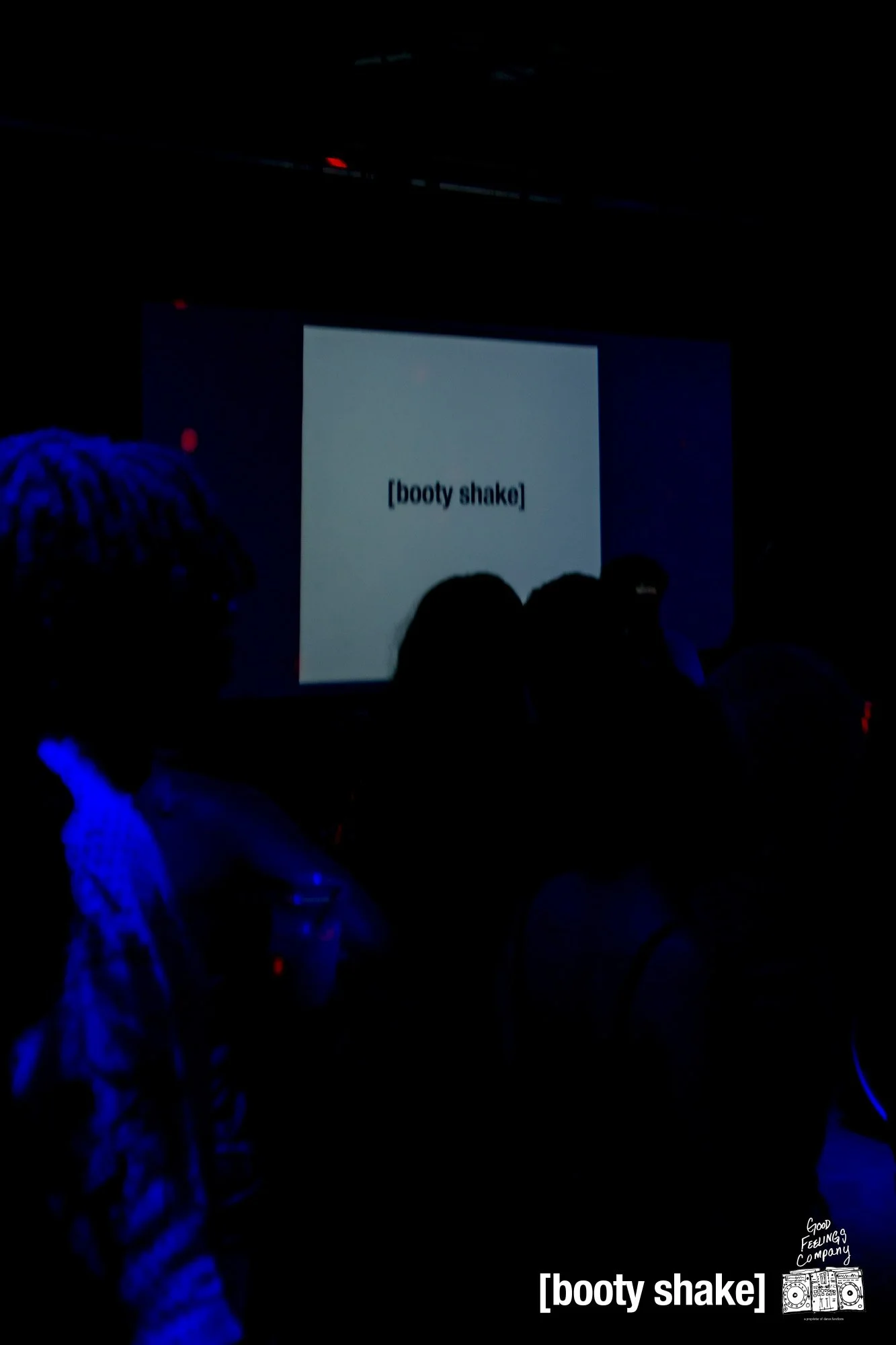 Dark room with a projection screen displaying the text '[booty shake]'. Silhouettes of people sitting and standing, some holding drinks. The scene suggests a party or event with a humorous or playful tone.