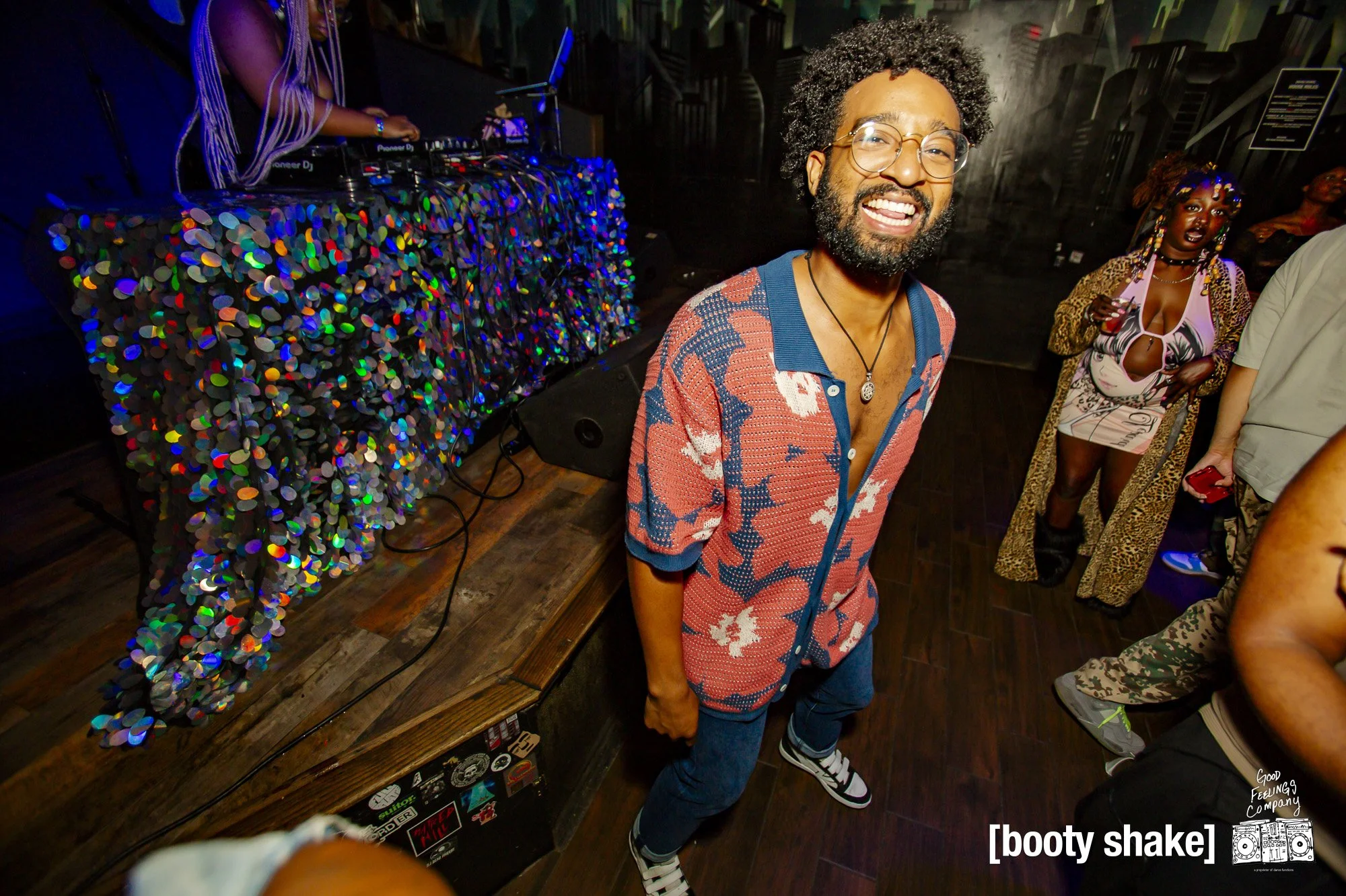 A man with glasses and a beard smiling at a party, wearing a patterned red and blue shirt and jeans. DJ equipment is visible in the background, and other partygoers are nearby, including a woman wearing a leopard-print coat and bold makeup. The capti