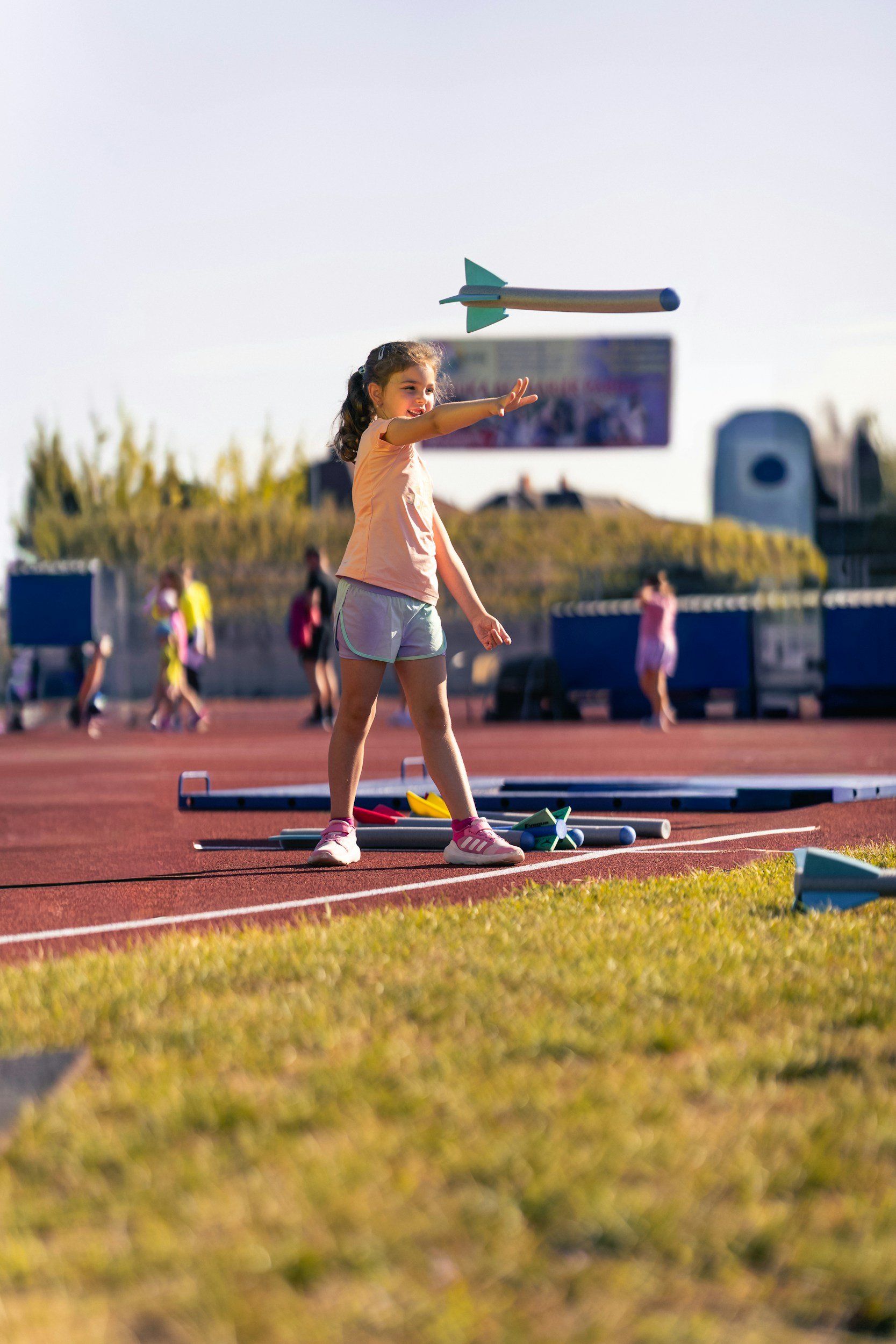 A young girl is throwing a javelin on a red athletic track, with several javelins on the ground nearby, and other people in the background.