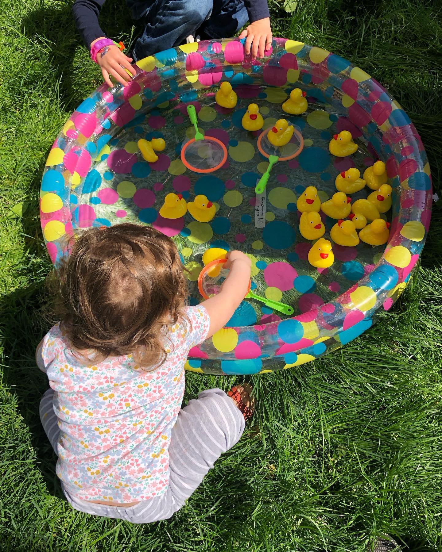 Child with curly hair sitting on green grass, playing with yellow rubber ducks in a small inflatable pool with colorful polka dots, using green and orange fishing nets.
