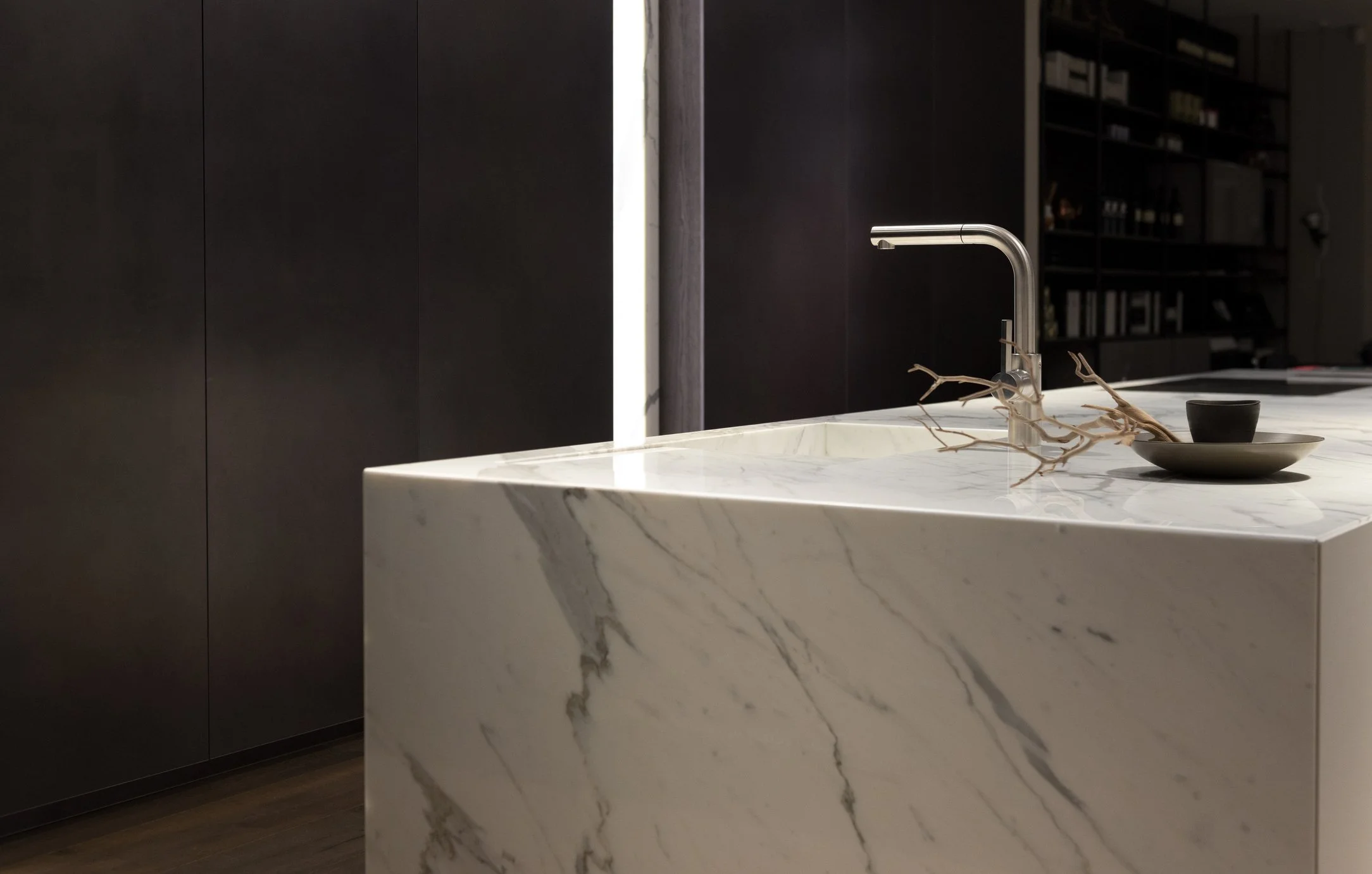 A kitchen countertop with a marble surface, a modern stainless steel faucet, and a small black bowl placed on a matching marble plate. In the background, there are dark cabinets and a bookshelf with various items.