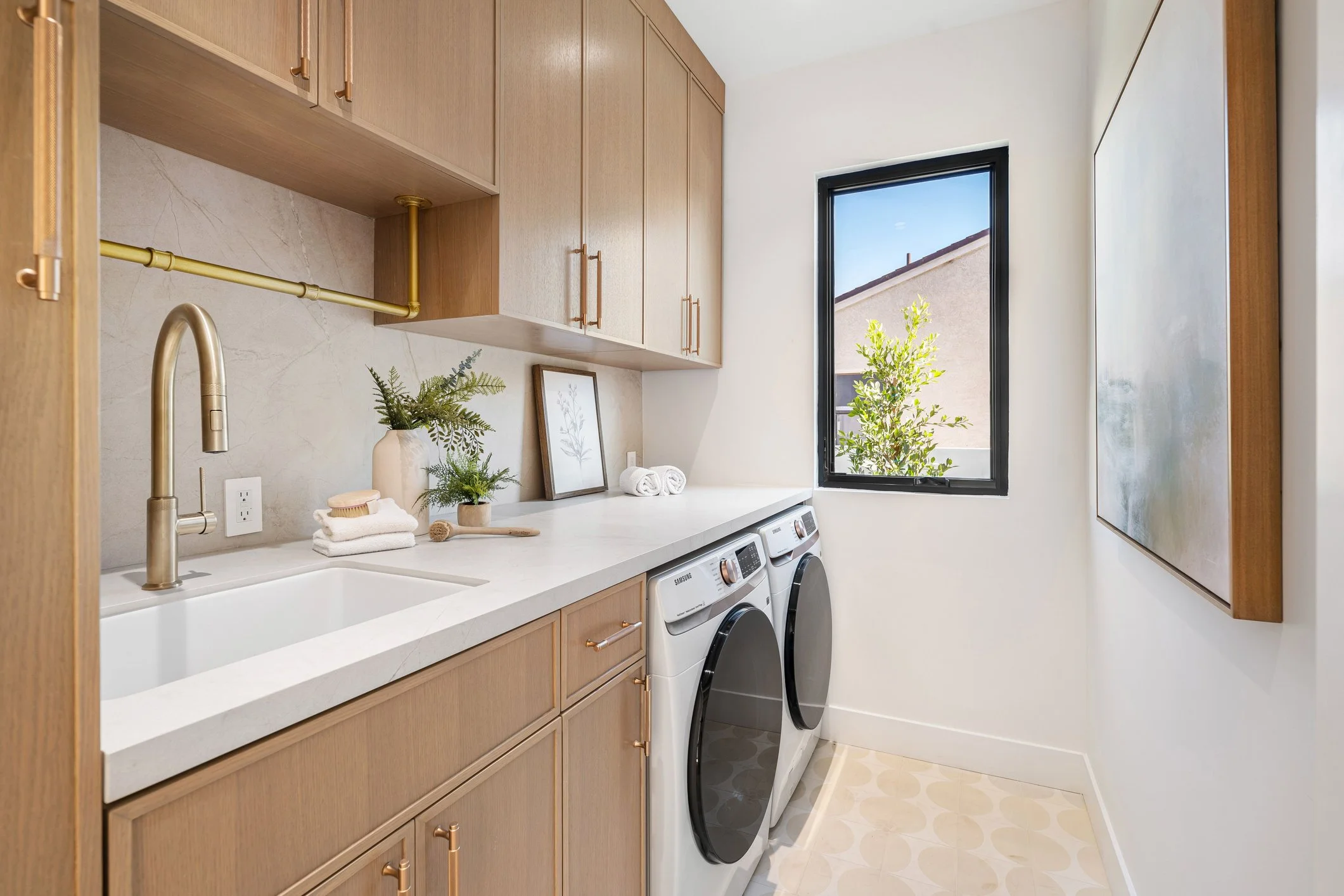 Laundry room with wooden cabinets, a white countertop, a gold pipe, a gold faucet, a framed picture, towels, plants, a window with a view of the roof and sky, and a washing machine and dryer.