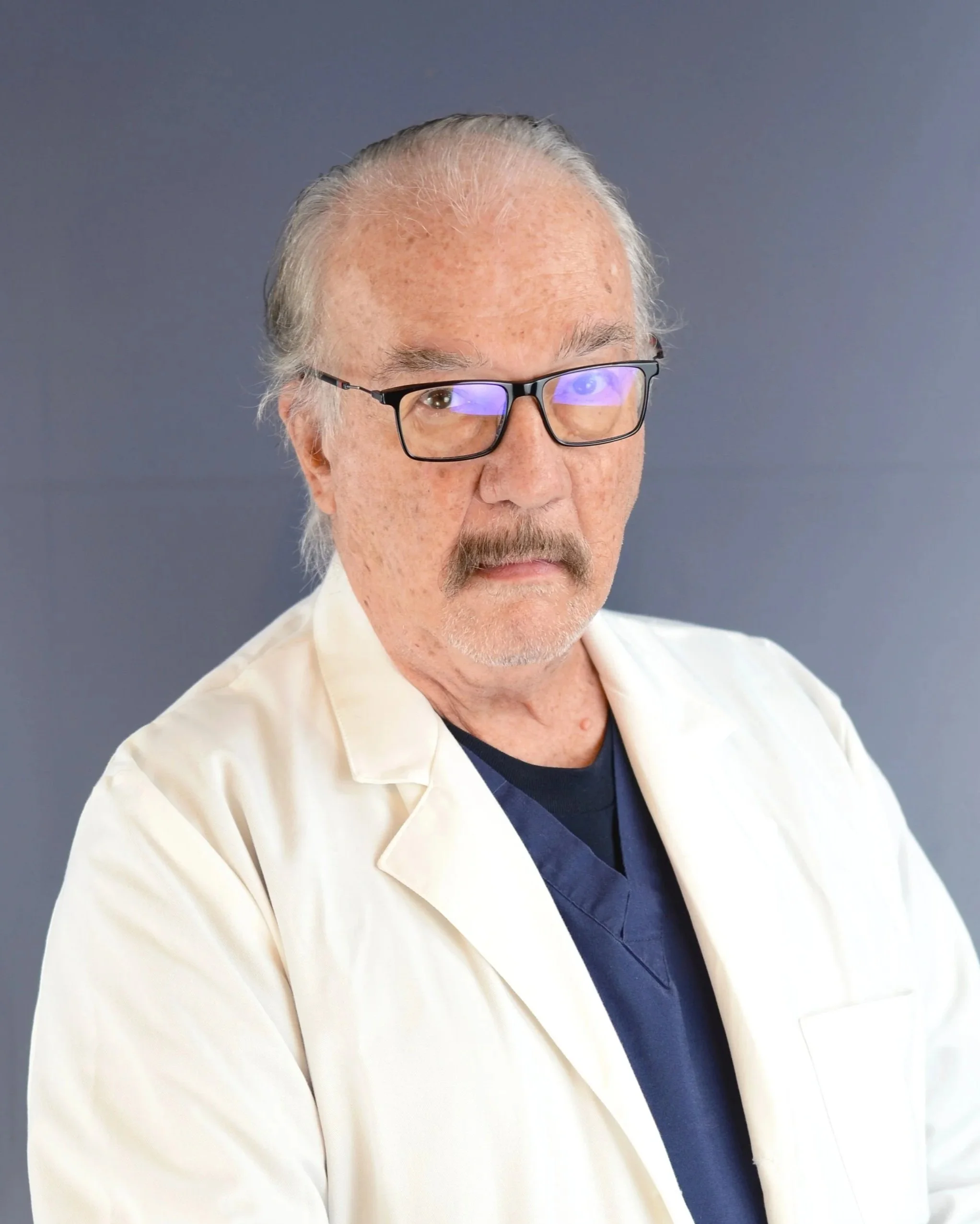 A senior male doctor or scientist wearing glasses, a white coat, and a navy scrubs shirt, standing against a gray background.