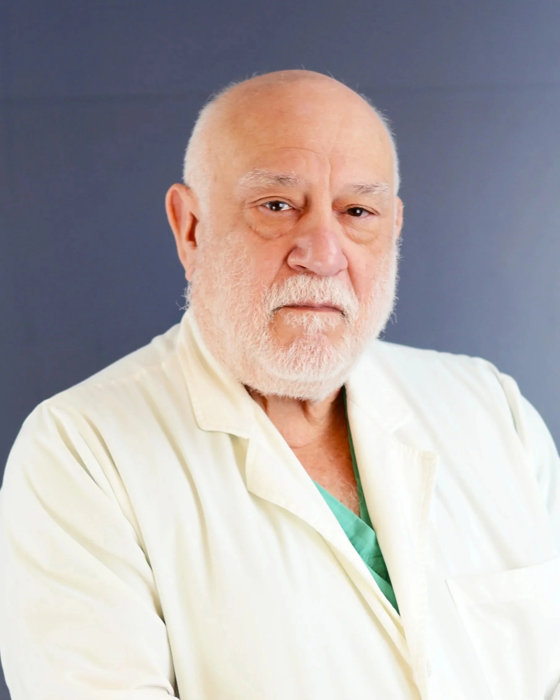 A senior man with a white beard and mustache, bald head, and wearing a white medical coat over green scrubs, standing against a plain gray background.