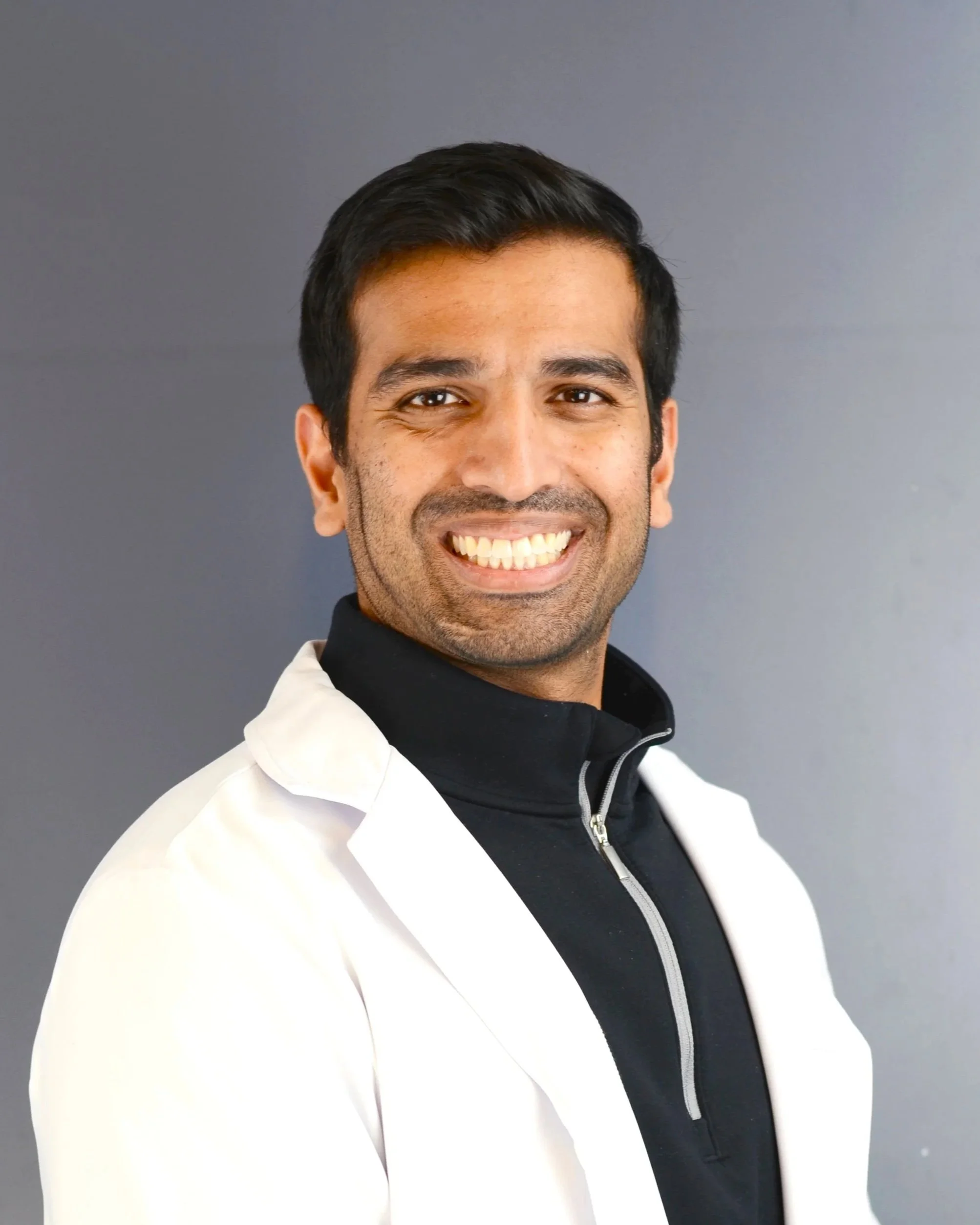 Portrait of a man with dark hair, smiling, wearing a black zip-up jacket and a white coat, standing against a gray background.