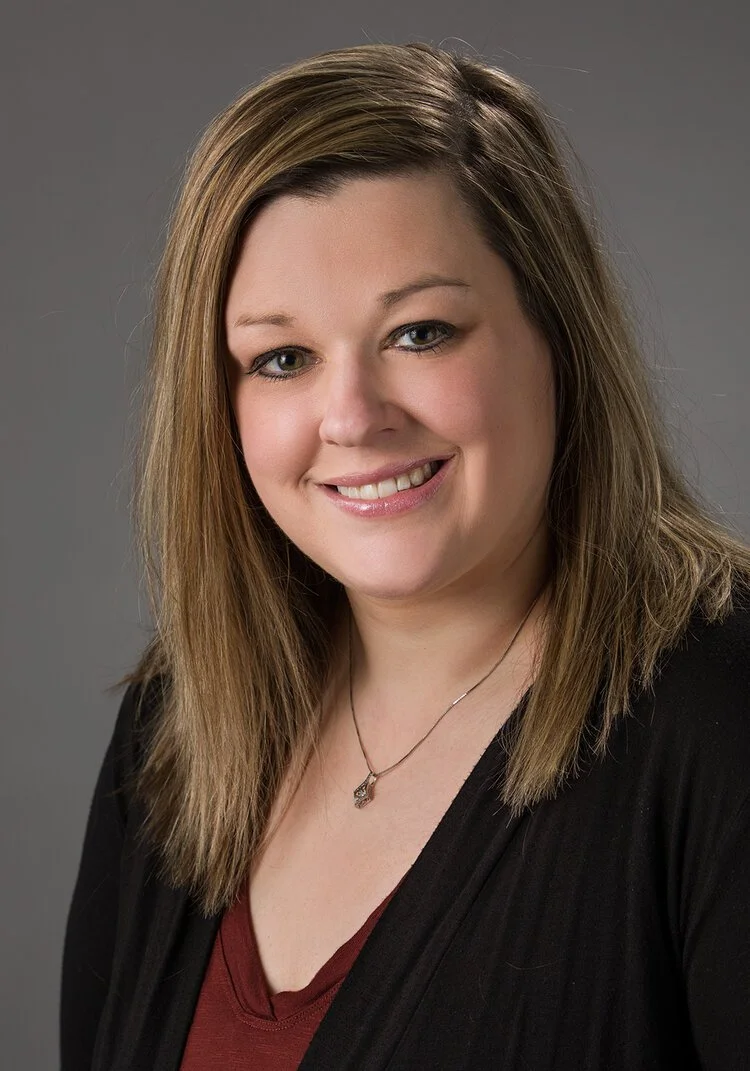 A woman with shoulder-length light brown hair wearing a black blazer and a maroon top, smiling at the camera against a gray background.