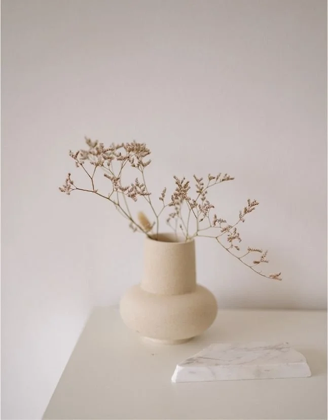 A beige ceramic vase with dried flowers on a white surface, with a small white marble slab nearby against a plain, light background.