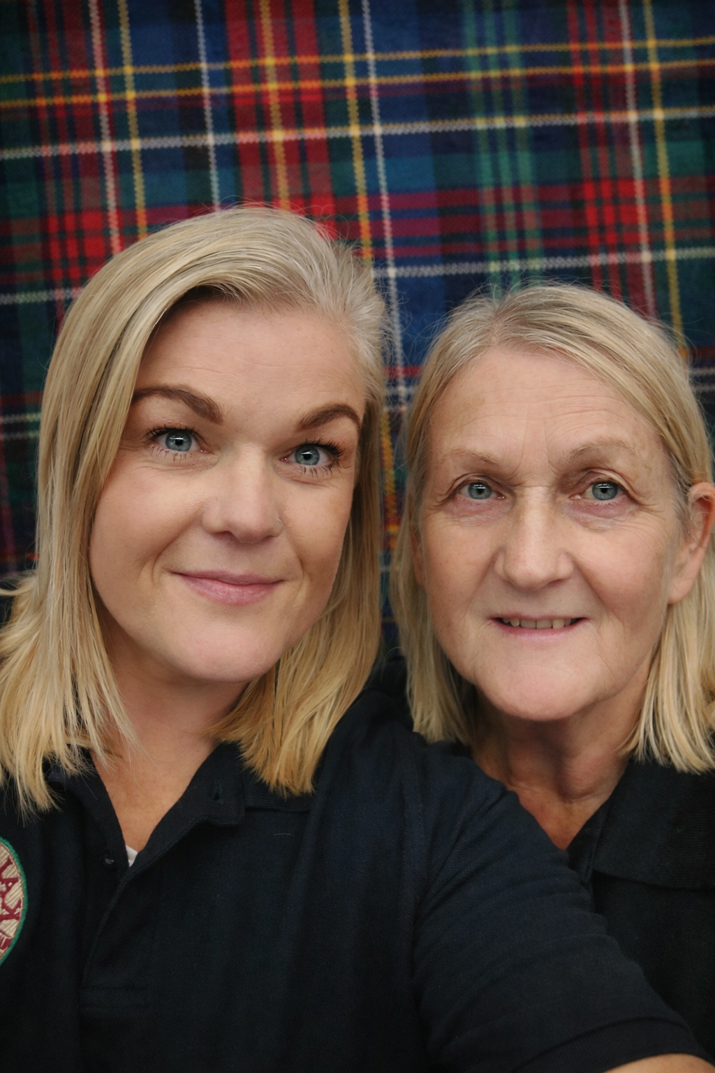 Two women with blonde hair and blue eyes taking a close-up selfie in front of a tartan patterned background.