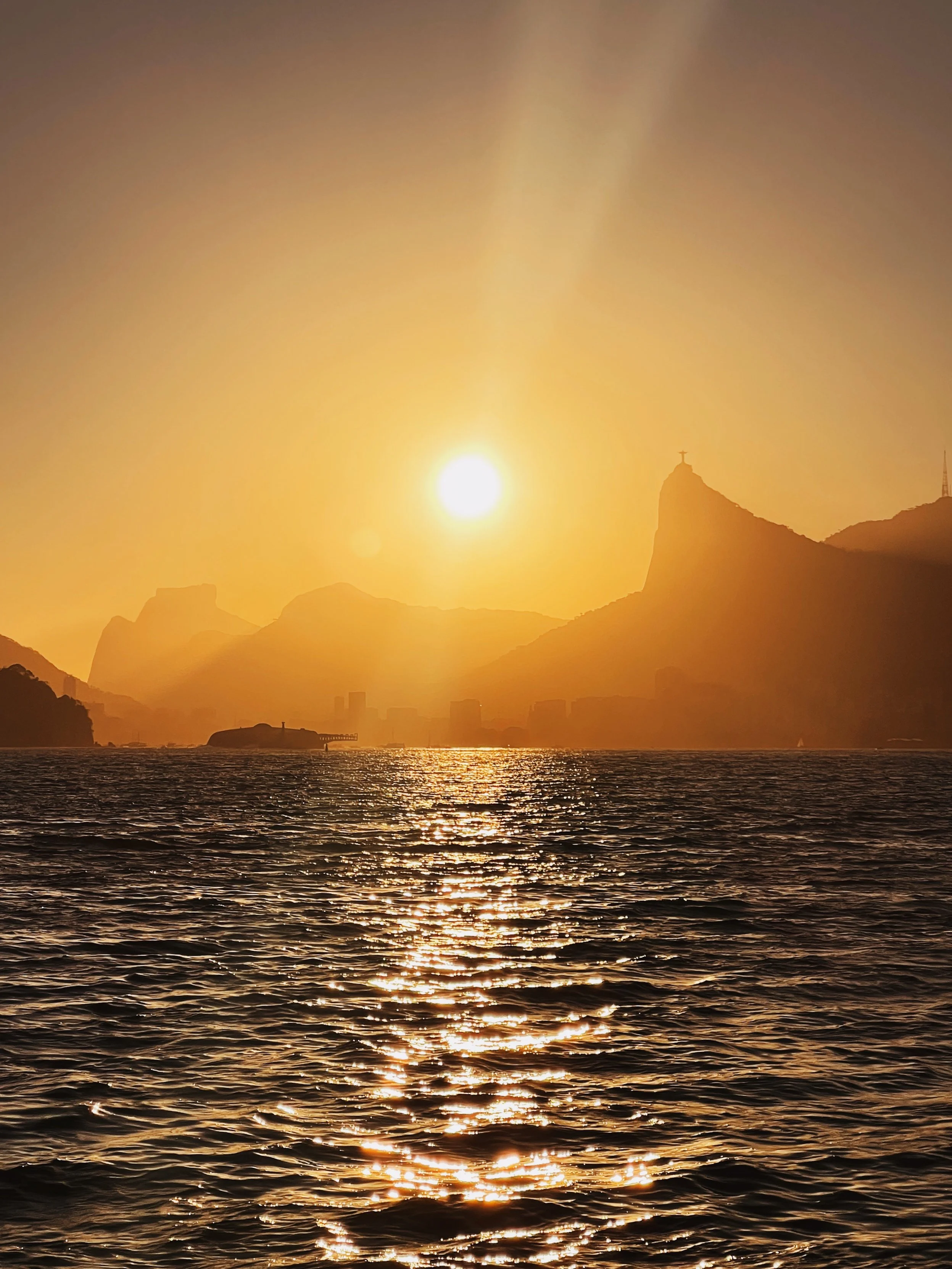 Pôr do sol visto de frente sobre a Baía de Guanabara, com o Cristo Redentor ao fundo na encosta da montanha.