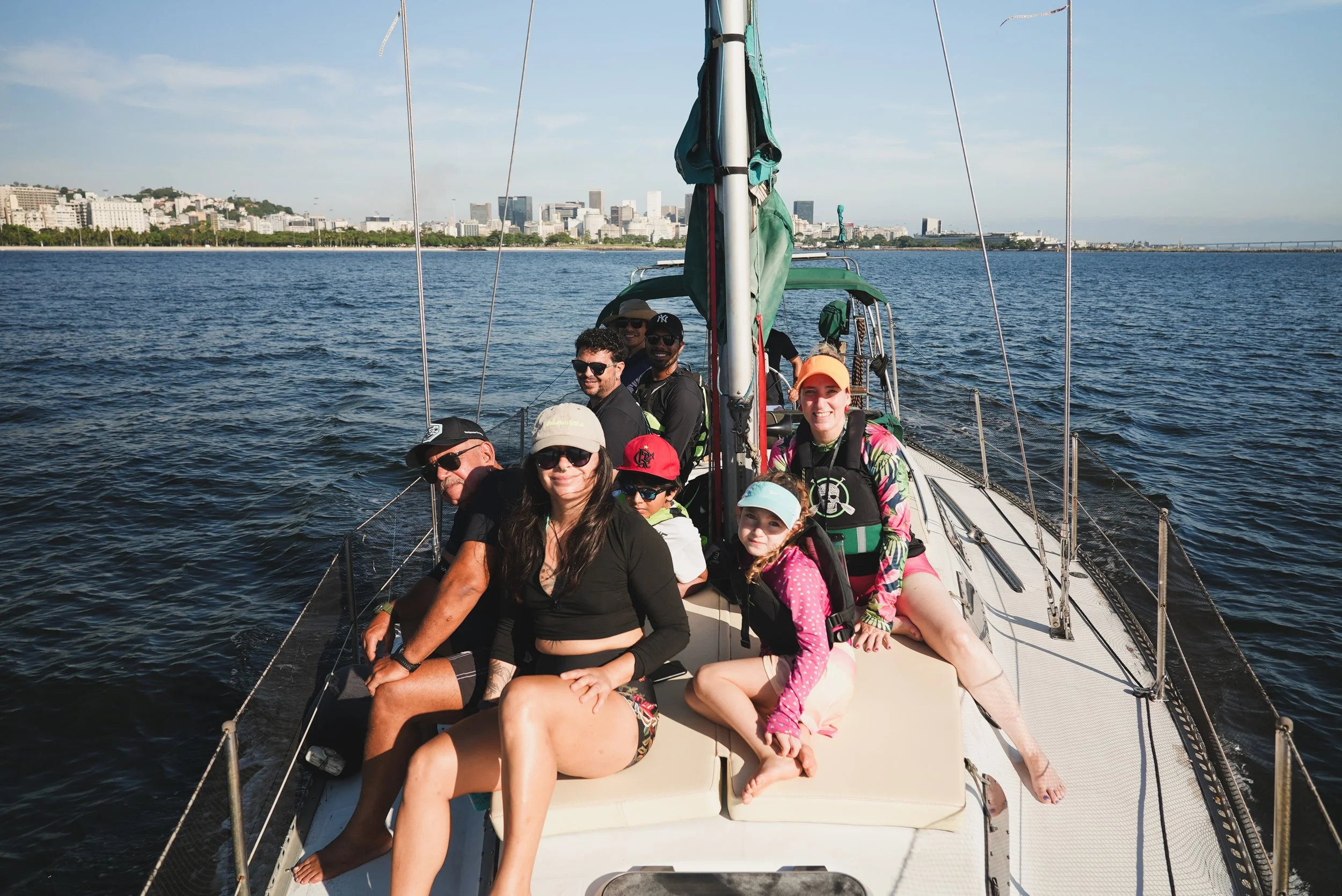 Pessoas sorrindo sentadas em um barco de vela no mar, com uma cidade ao fundo, em um dia ensolarado.