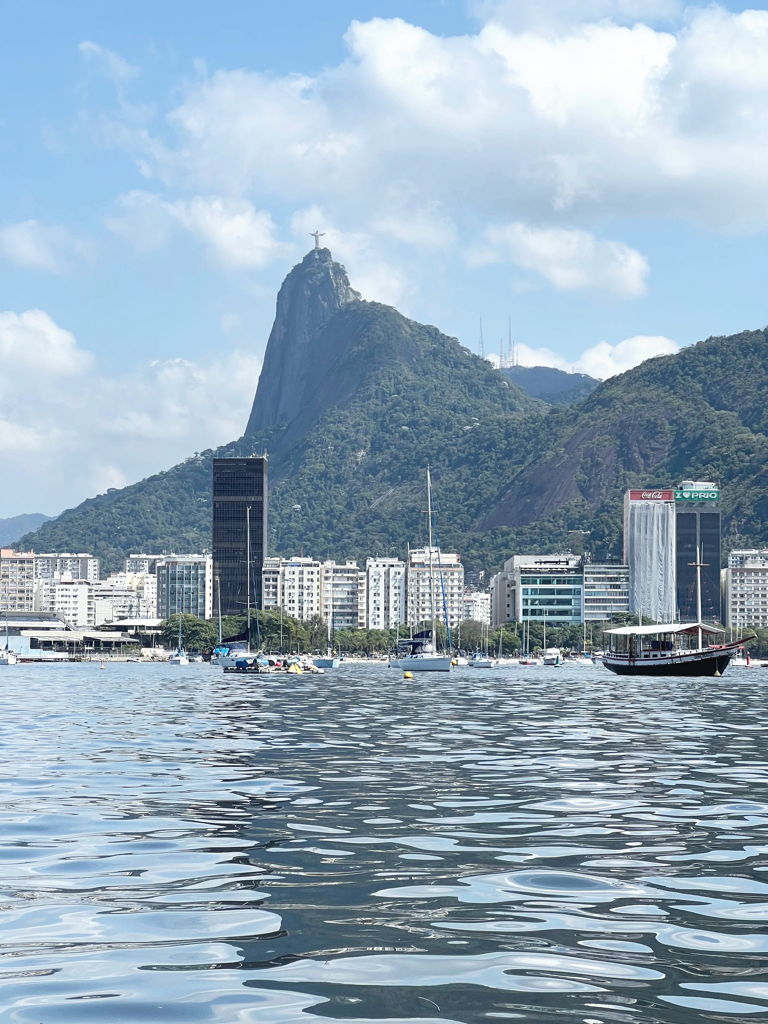 Paisagem de uma cidade costeira com o Cristo Redentor ao fundo, montanhas verdes, edifícios altos, barcos no rio e céu parcialmente nublado.