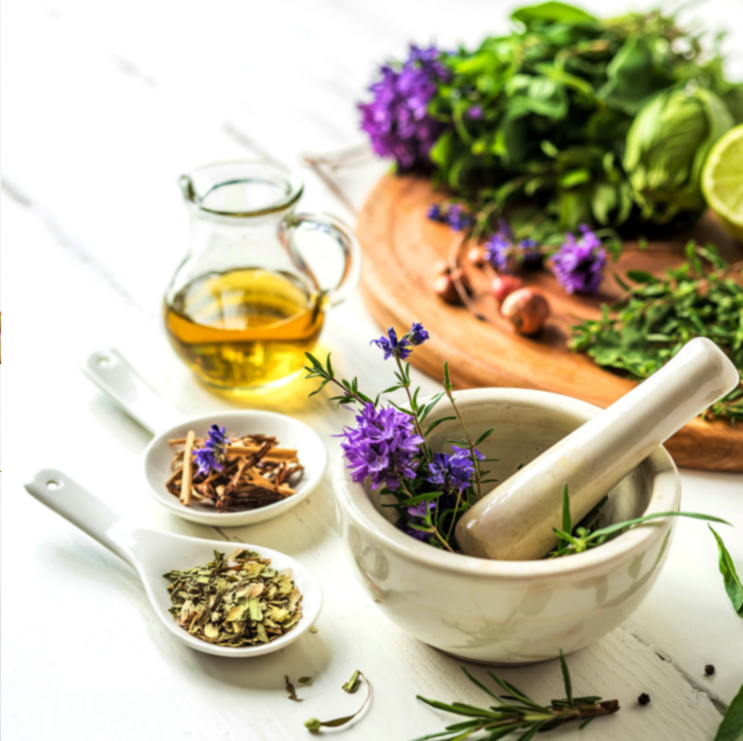 A white mortar and pestle with purple flowers inside, surrounded by small bowls of dried herbs, a glass jar of olive oil, and fresh herbs on a wooden cutting board.