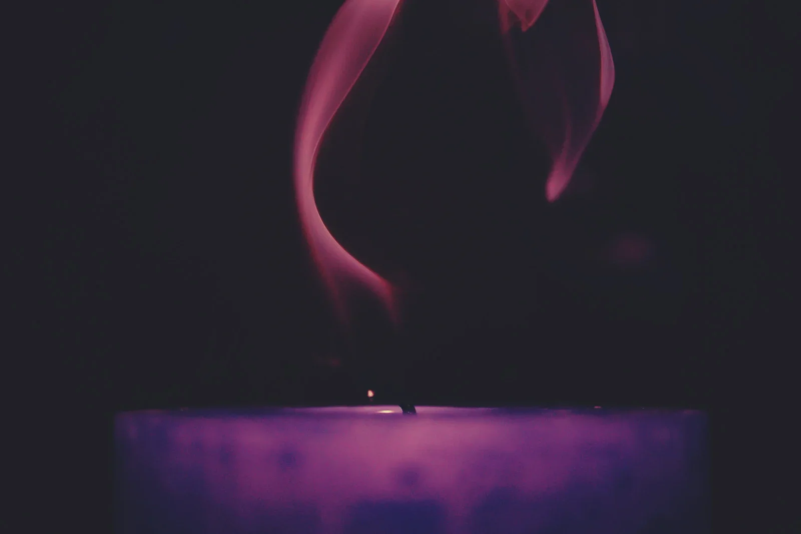 Close-up of a purple candle with pink smoke rising from its wick in darkness.