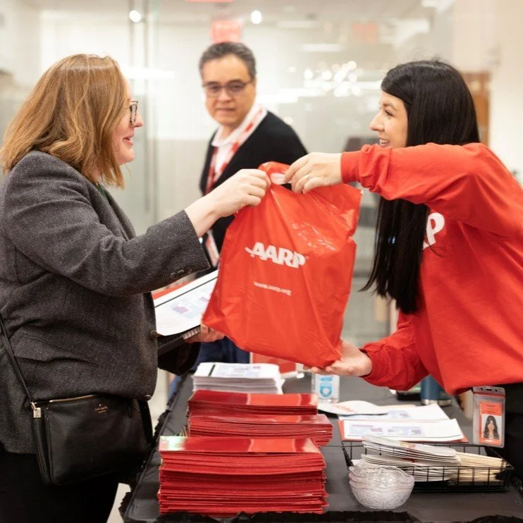 A woman is receiving a red AARP bag from a representative behind a table with informational materials, at an event. Another man is in the background.