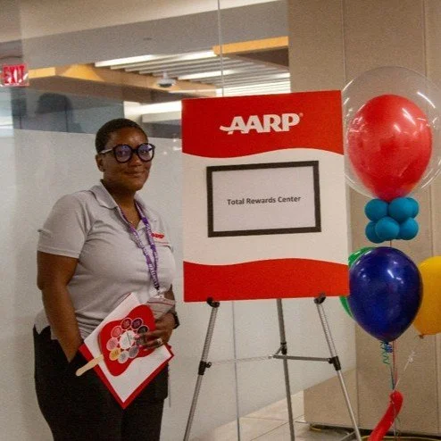 A woman standing next to an AARP Total Rewards Center sign with colorful balloons on the right side, holding a red folder with AARP logo, inside a building.