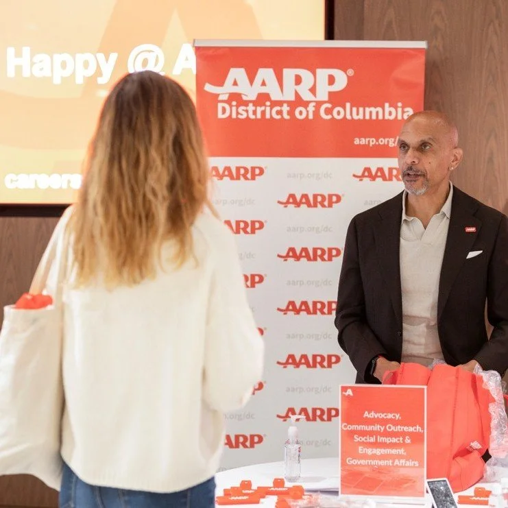 A man in a suit talking to a woman at an AARP event with a red and white banner that reads 'AARP District of Columbia.' There are promotional materials on the table, including a sign highlighting advocacy, community outreach, social impact, engagement, and government affairs.