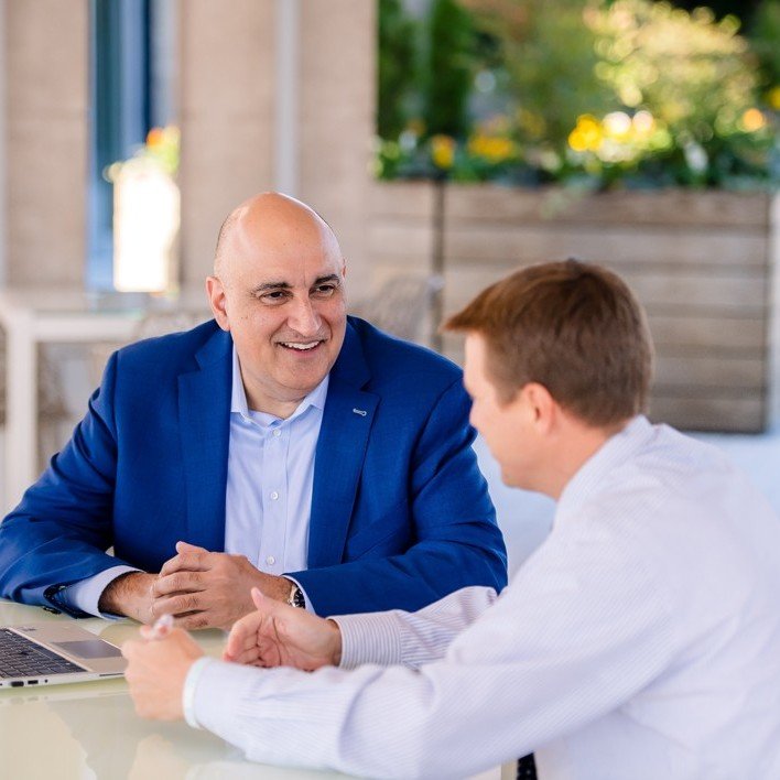 Two men engaged in conversation at a table, one in a blue suit and the other in a white shirt, in a professional setting with greenery in the background.
