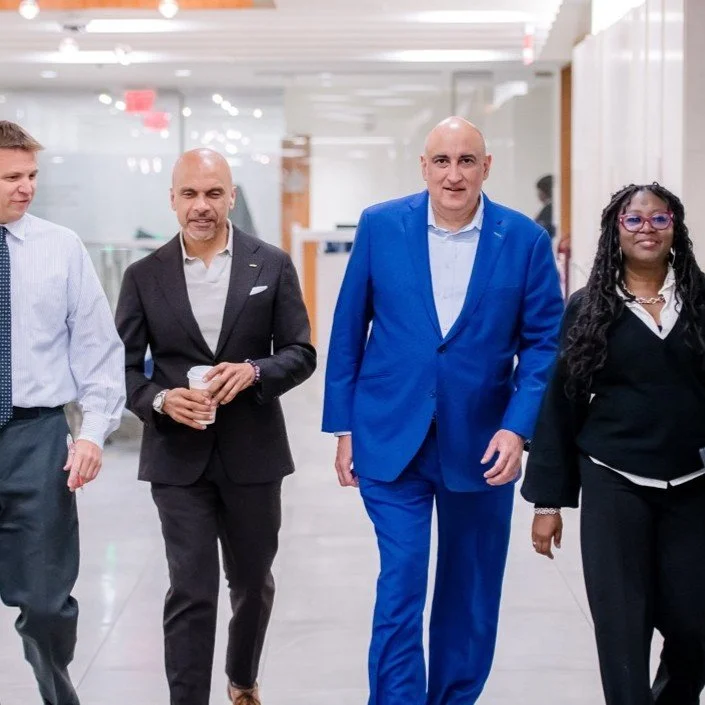 Four professionals walking in an office corridor, dressed in business attire, with bright lighting and modern decor.