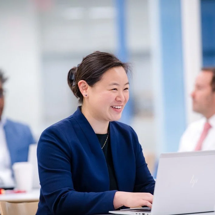 A woman smiling while working on a laptop in an office setting with other business professionals in the background.