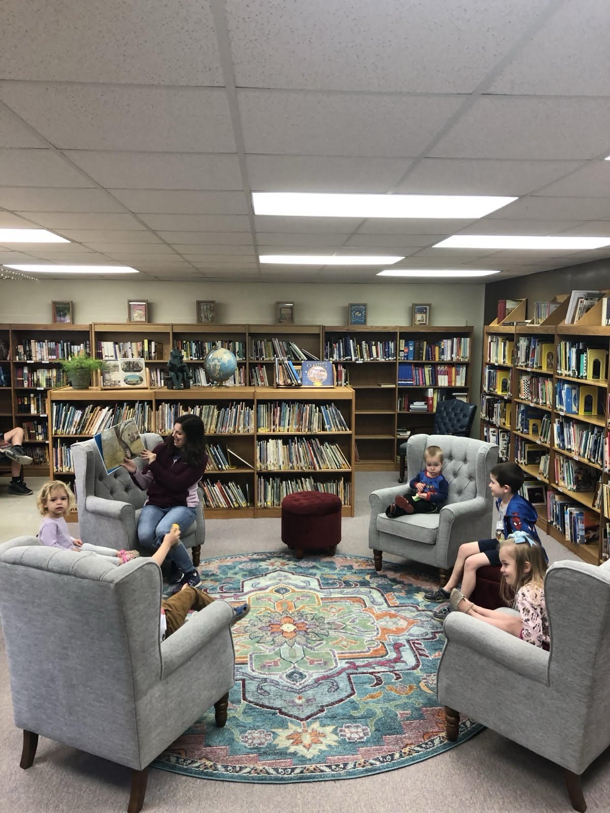 Sadie, a board member, reading a book to children in a library with bookshelves and a colorful rug.