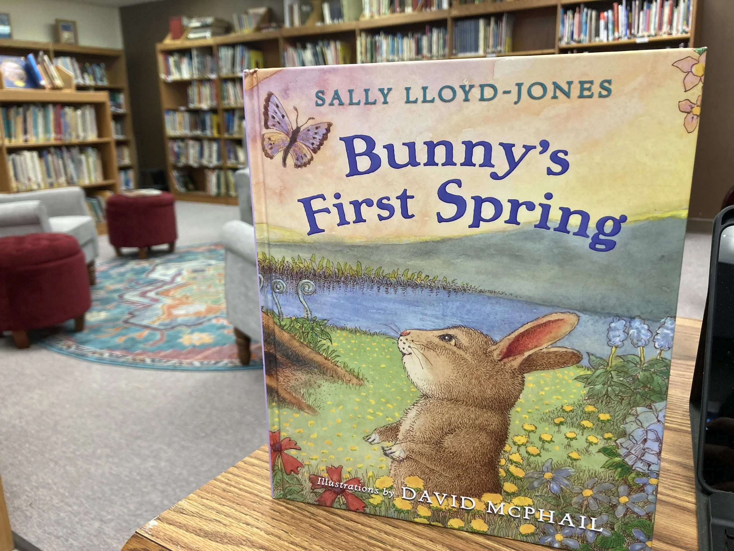 an image of the book Bunny's First Spring sitting on top of the desk in the Stinesville Community Library. Bookshelves filled with childrens books in the background and cozy chairs to sit in.