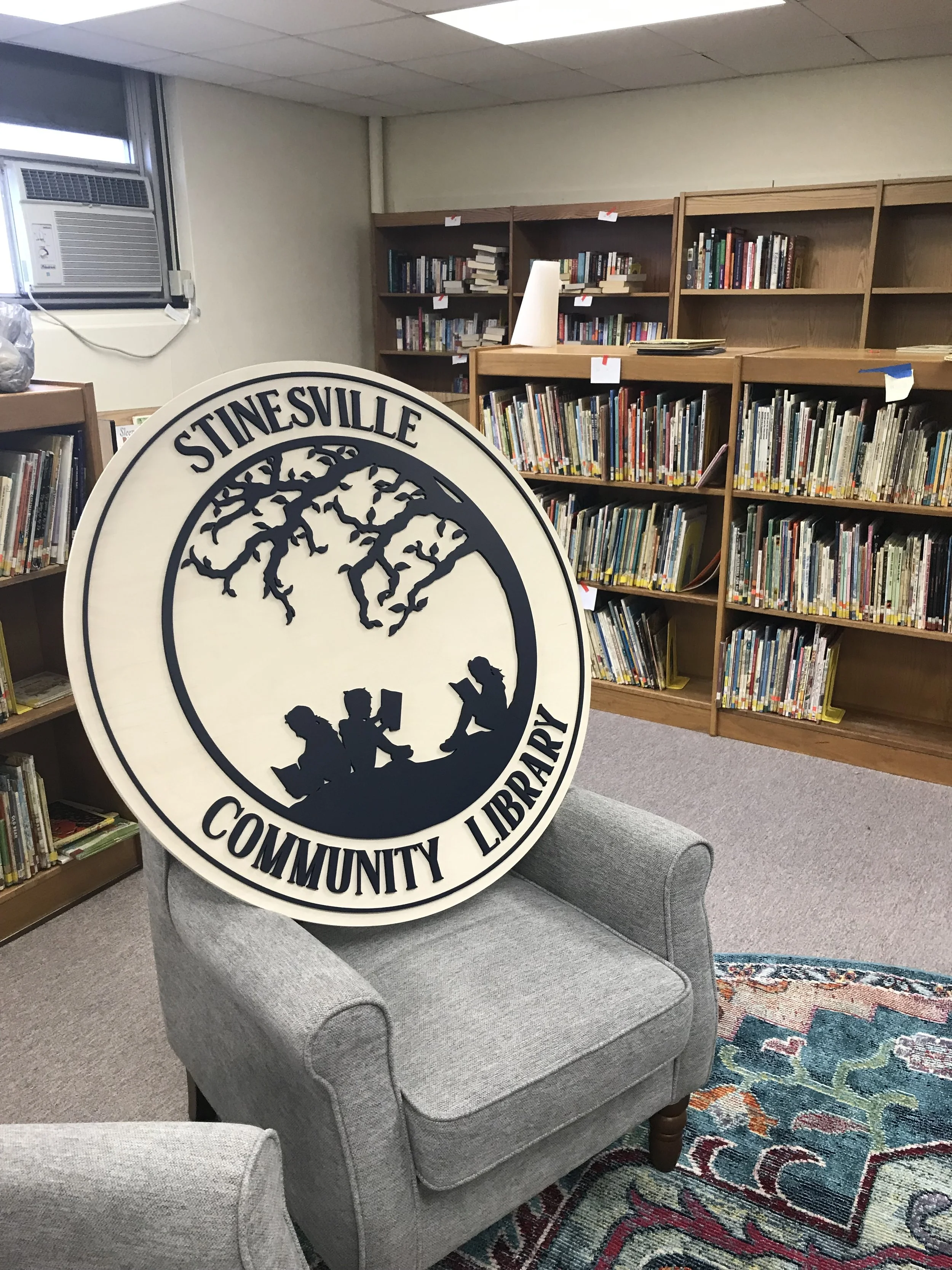 A circular sign for Stinesville Community Library showing its logo of a silhouette of three children reading underneath a tree.