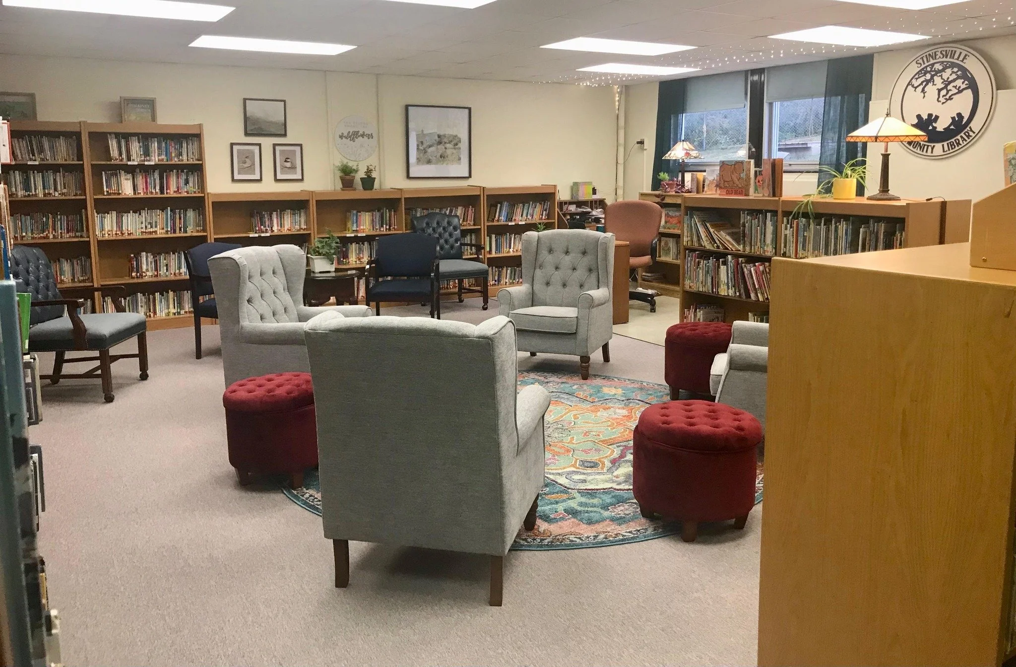 A cozy library reading area with armchairs, ottomans, and bookshelves, illuminated by ceiling and table lamps.