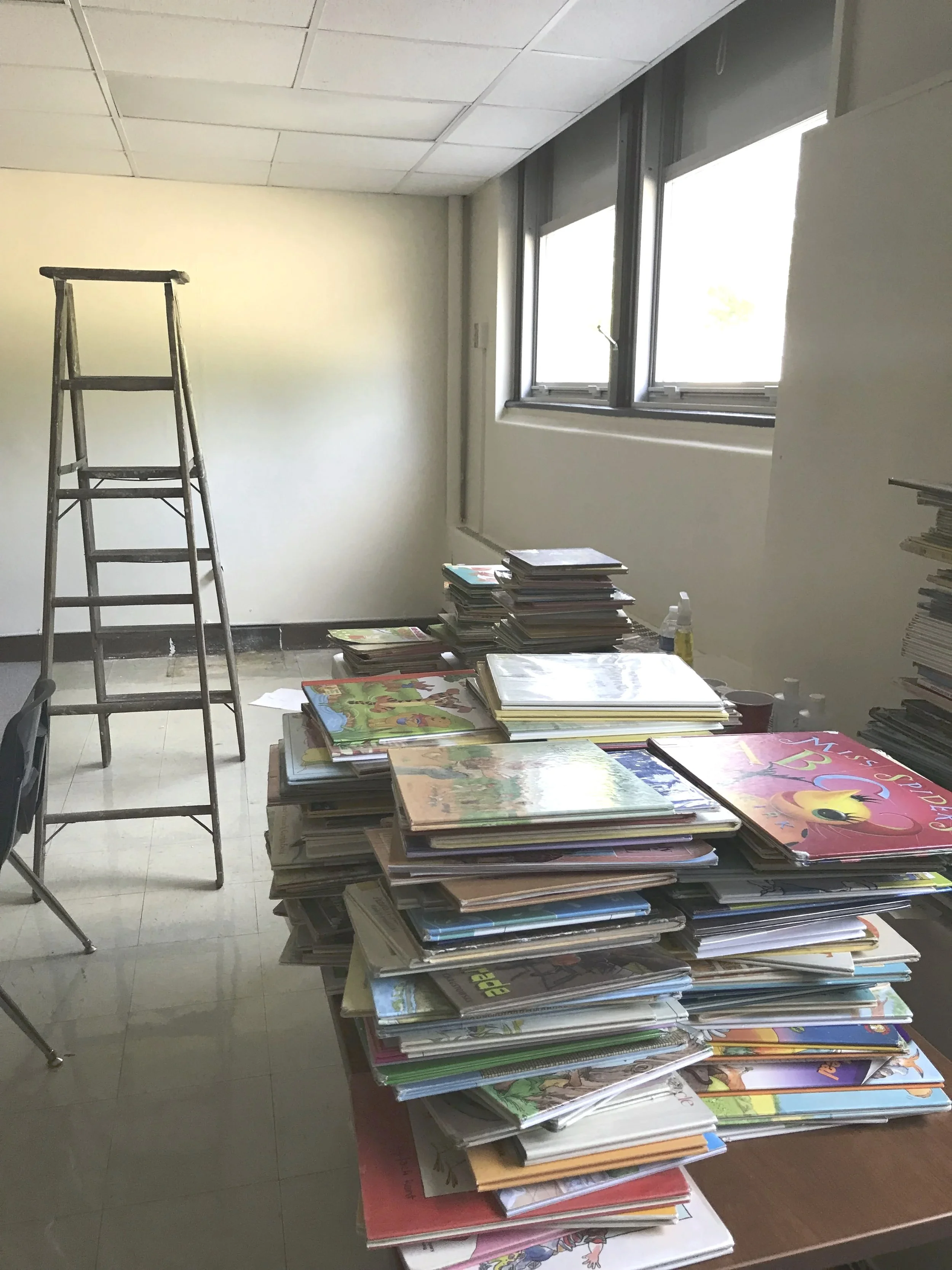 A room with a large pile of children's books and stack of magazines on a table, a ladder, and a window with sunlight shining through showing the library under construction.