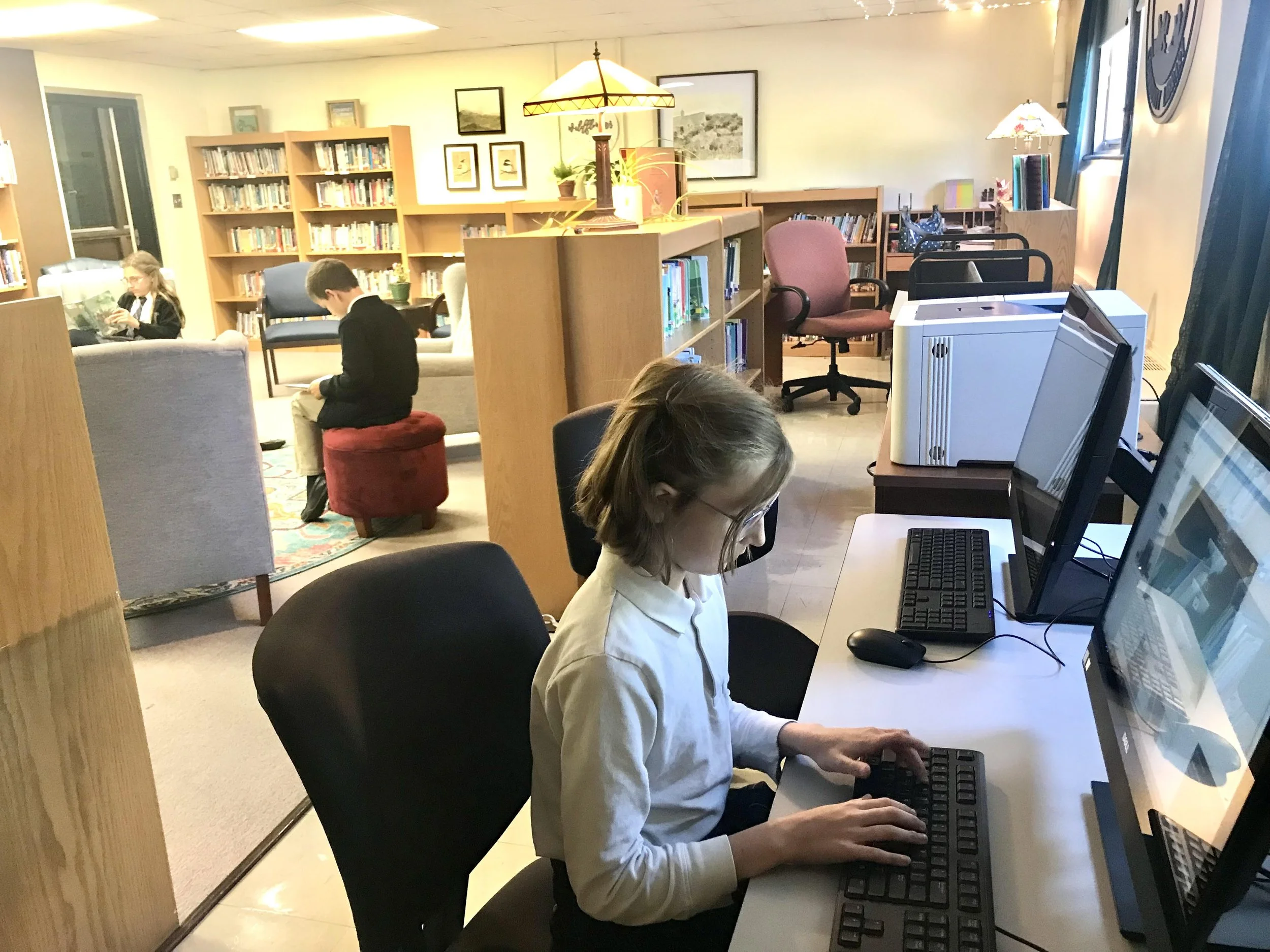 A girl is working on a computer at a desk in a library, with bookshelves, other children, and a reading area visible in the background.