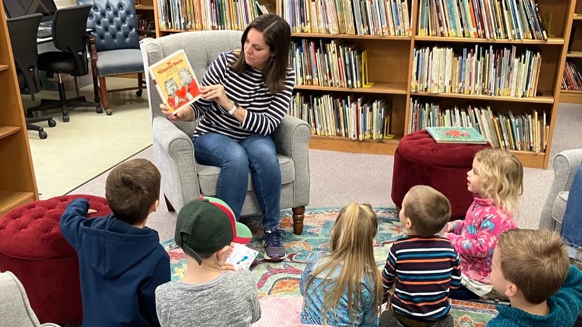 A woman reading a children's book titled 'Valentine Bear' to a group of six children in a library, with bookshelves in the background.