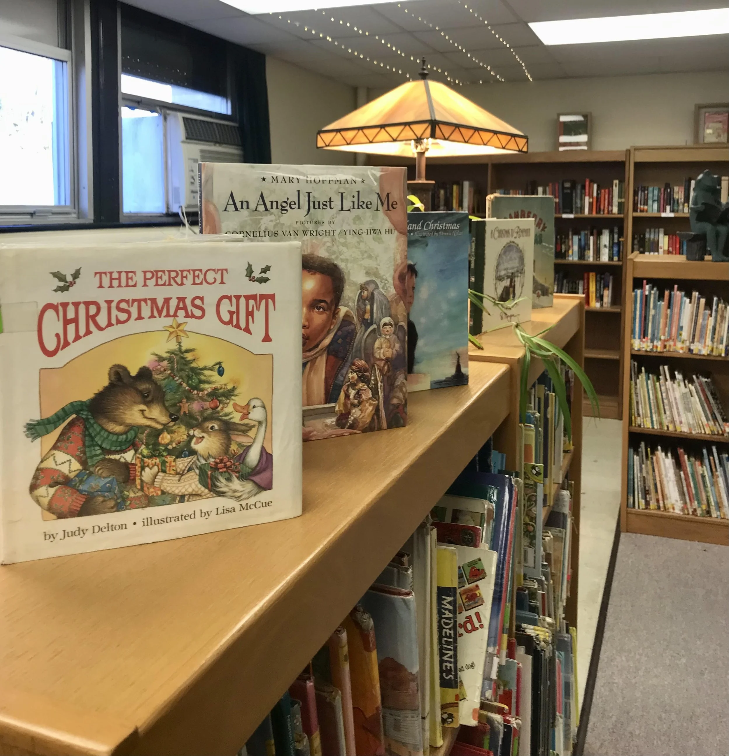 A display of children's books on a wooden shelf in a library, including titles such as "The Perfect Christmas Gift" and "An Angel Just Like Me," with books arranged on a tilted surface and additional books on shelves in the background.