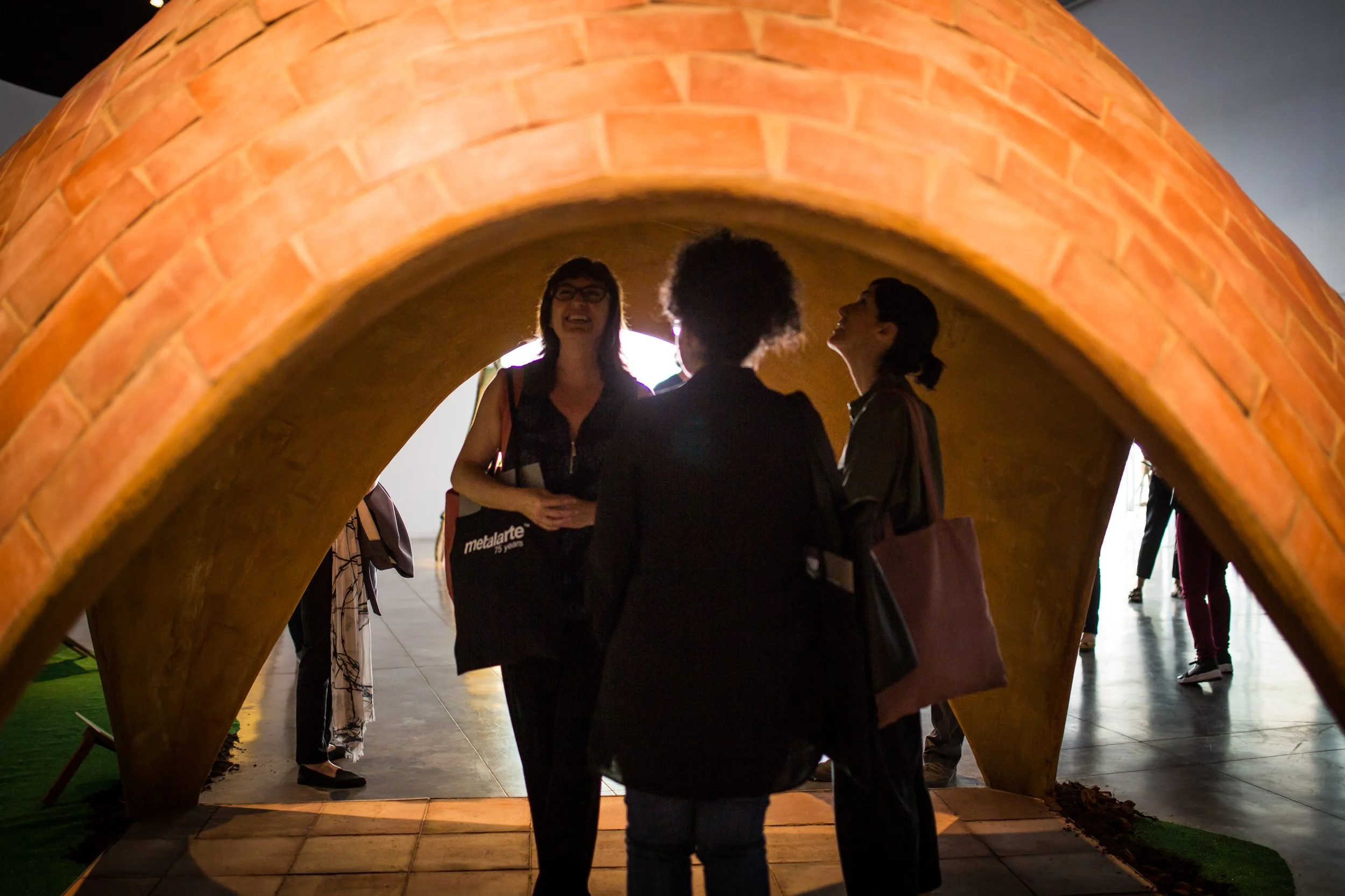 Three women are seen from behind and inside a arch-like structure with a brick exterior, engaging in conversation at an indoor Musuem event.