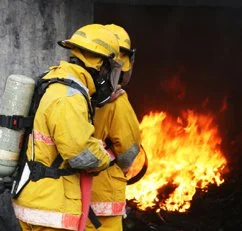 Two firefighters in yellow gear and helmets standing near a large fire.