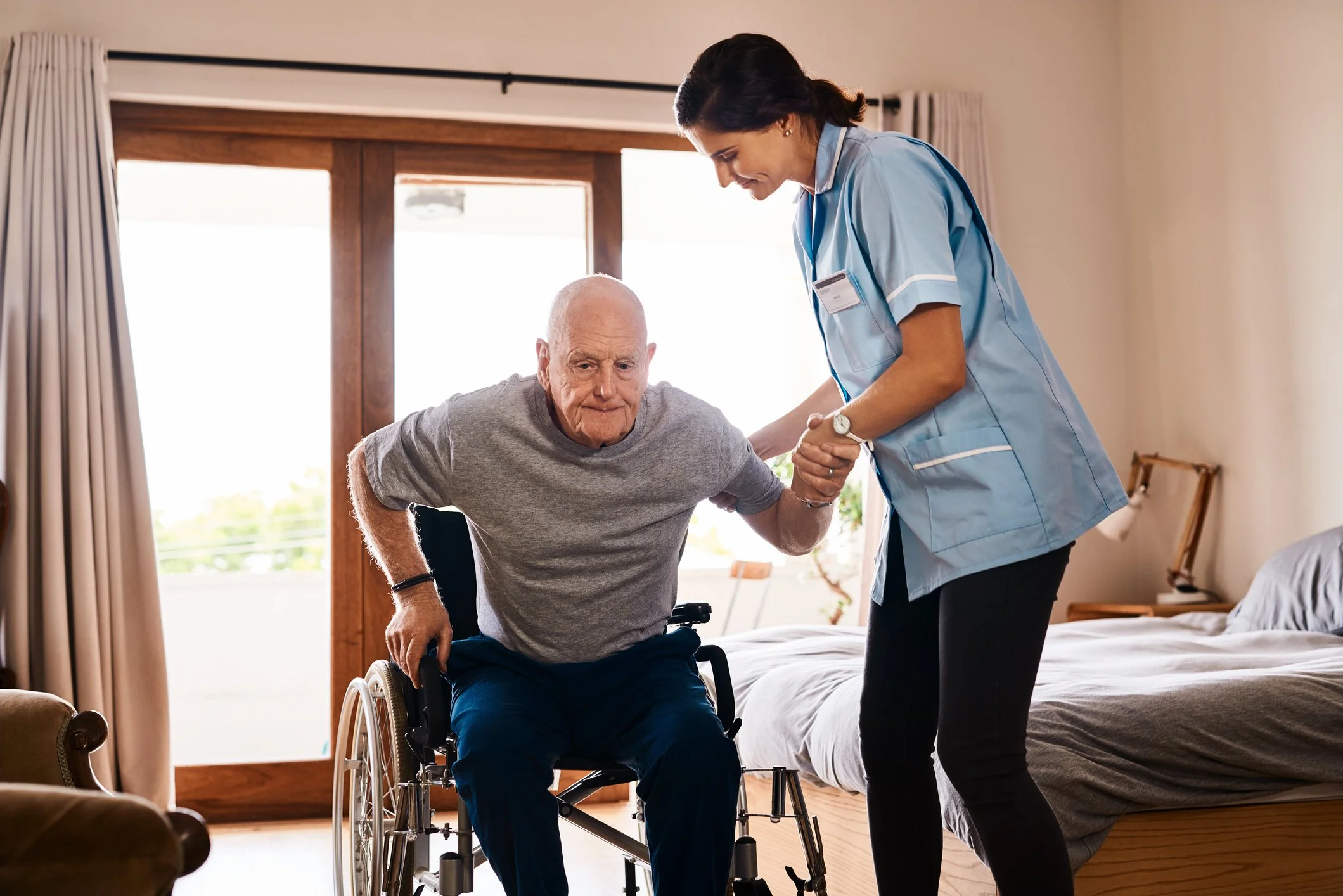 A caregiver helping an elderly man in a wheelchair to stand in a brightly lit room with large windows and curtains.