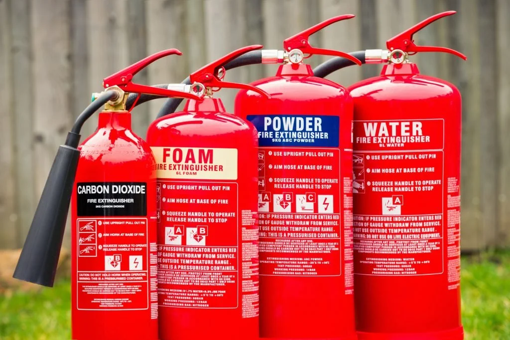 Four red fire extinguishers labeled for water, foam, carbon dioxide, and powder, with hoses and gauges, outdoors on a grassy area with a wooden fence in the background.