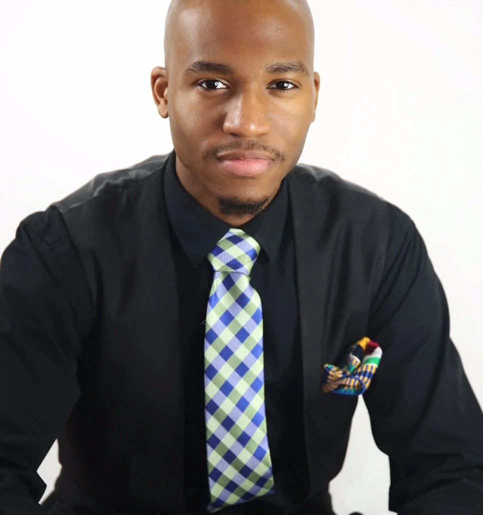 A young man with a shaved head and goatee, wearing a black shirt with a colorful patterned pocket square, and a plaid tie, posed against a plain white background.