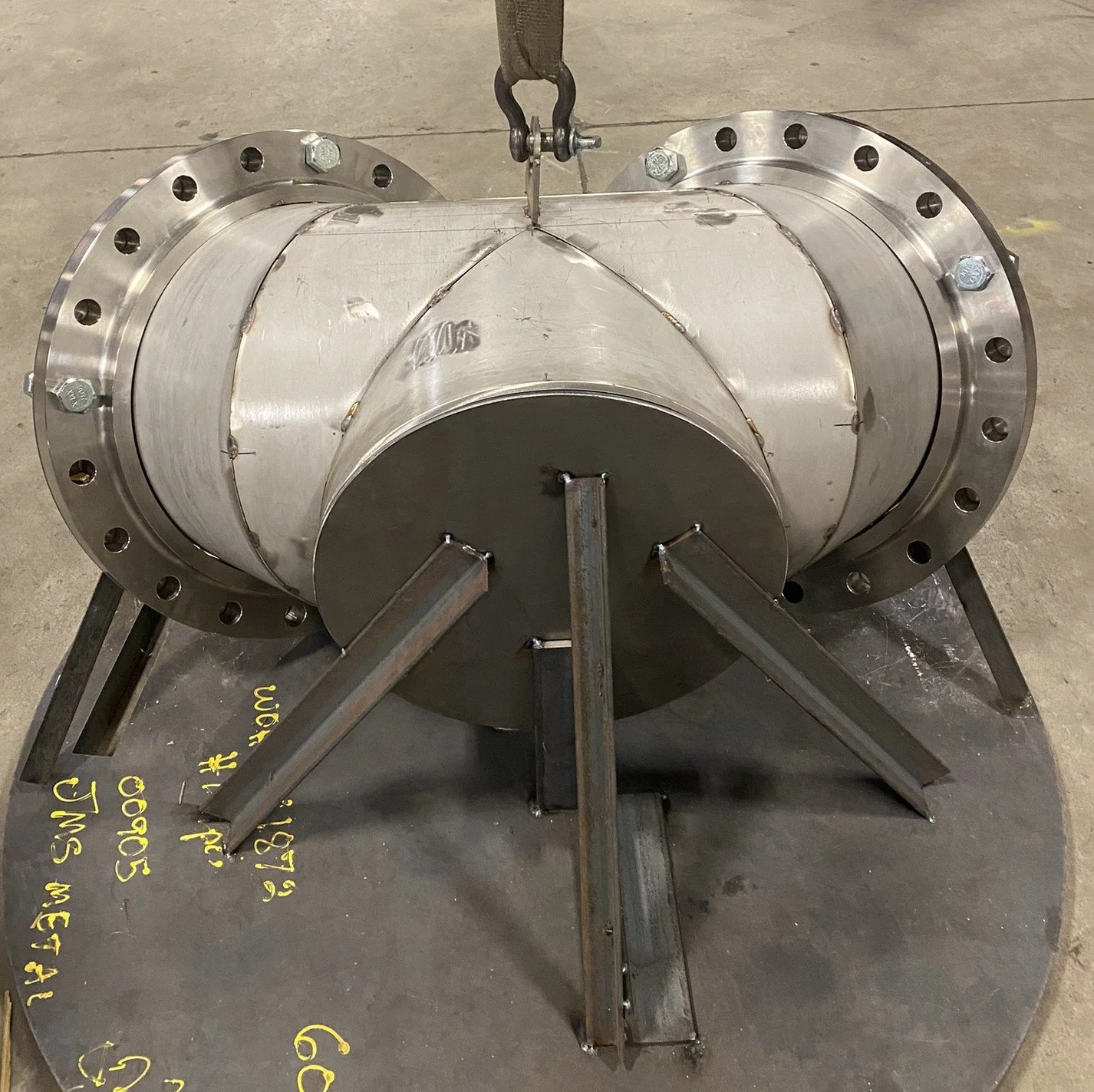 A large metal pressure vessel or pipe component with flanged ends, supported by welded steel stands, sitting on a metal plate with yellow writing on the floor.