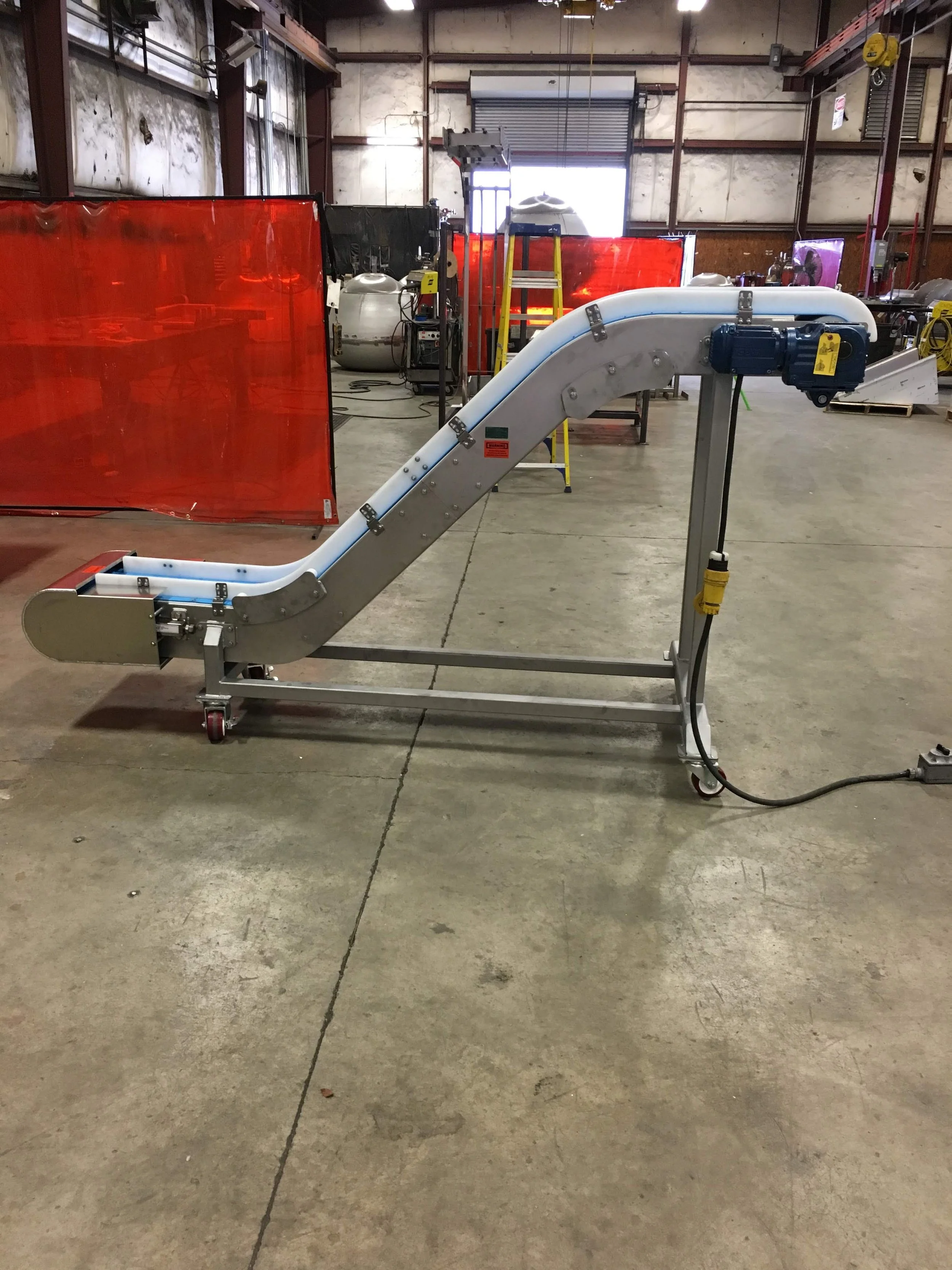A medical conveyor belt in a warehouse or industrial setting with a metal frame, white plastic belt, and a blue motor, surrounded by equipment, yellow safety barriers, and red privacy screens.