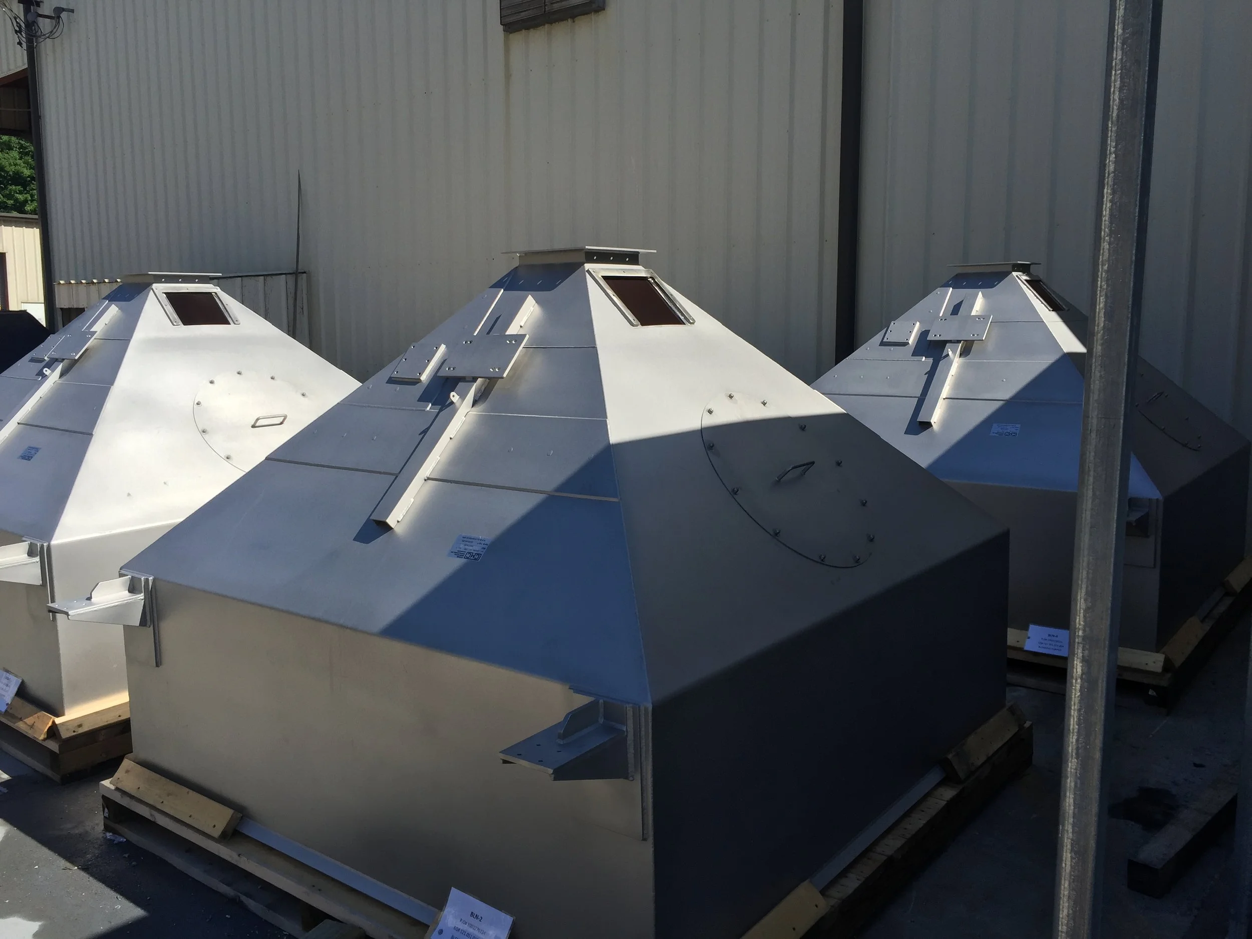 Three large metallic ventilation units with angular, pyramid-like shapes are placed outdoors next to a metal building wall.